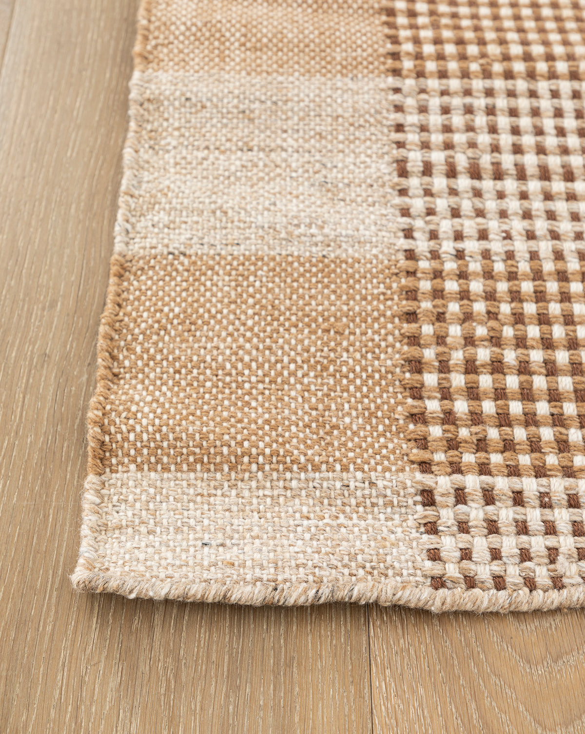 A close-up of the Hargrove Handwoven Indoor/Outdoor Rug, featuring a beige, cream, and brown checker pattern, shown on a light wooden floor.