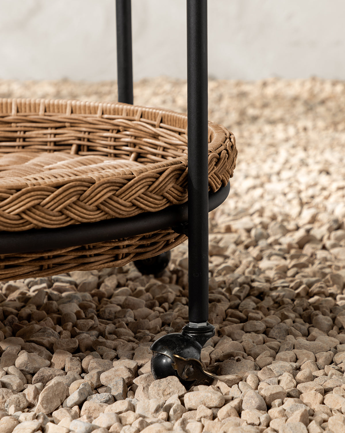 A close-up of the Haviland Bar Cart featuring weather-resistant wicker, a black metal frame, and caster wheels, set on a bed of small, light-colored pebbles, by McGee & Co.