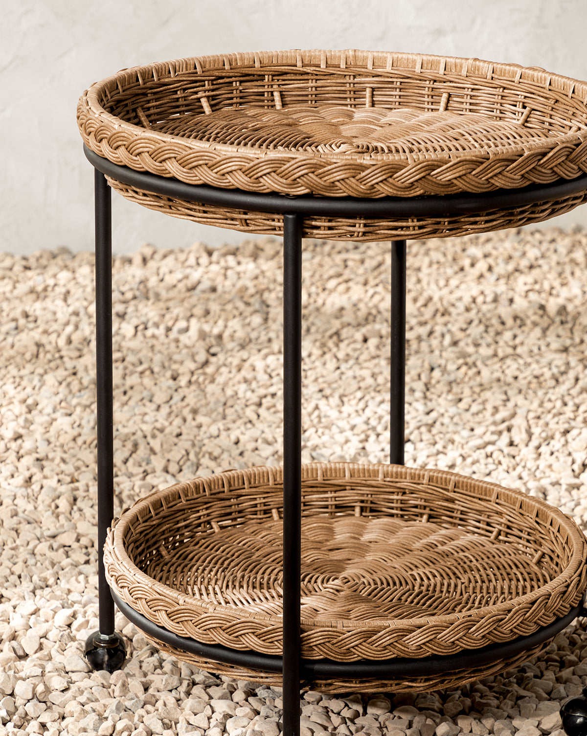 The Haviland Bar Cart features weather-resistant wicker with two round tiers and sturdy black metal legs, set on small white stones against a light textured wall—ideal for outdoor entertaining, by McGee & Co.