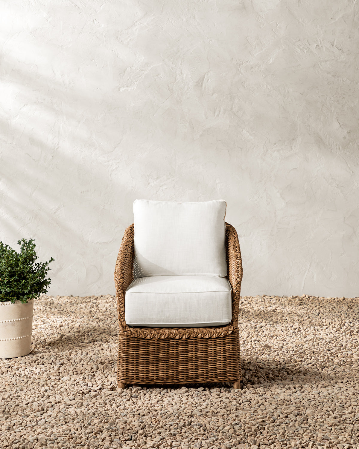 The Haviland Outdoor Dining Chair, featuring weather-resistant wicker and white cushions, is placed on gravel beside a potted green plant against a textured off-white wall, by McGee & Co.