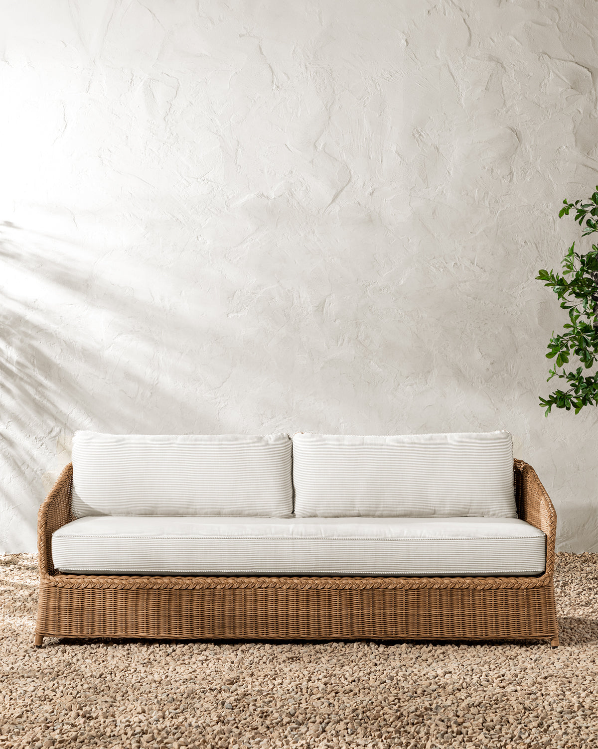 A Haviland Outdoor Sofa with Perennials Taupe Stripe 88 sits on a textured rug against a white, stucco-textured wall, with a green plant to the right and soft sunlight casting gentle shadows from the left by McGee & Co.