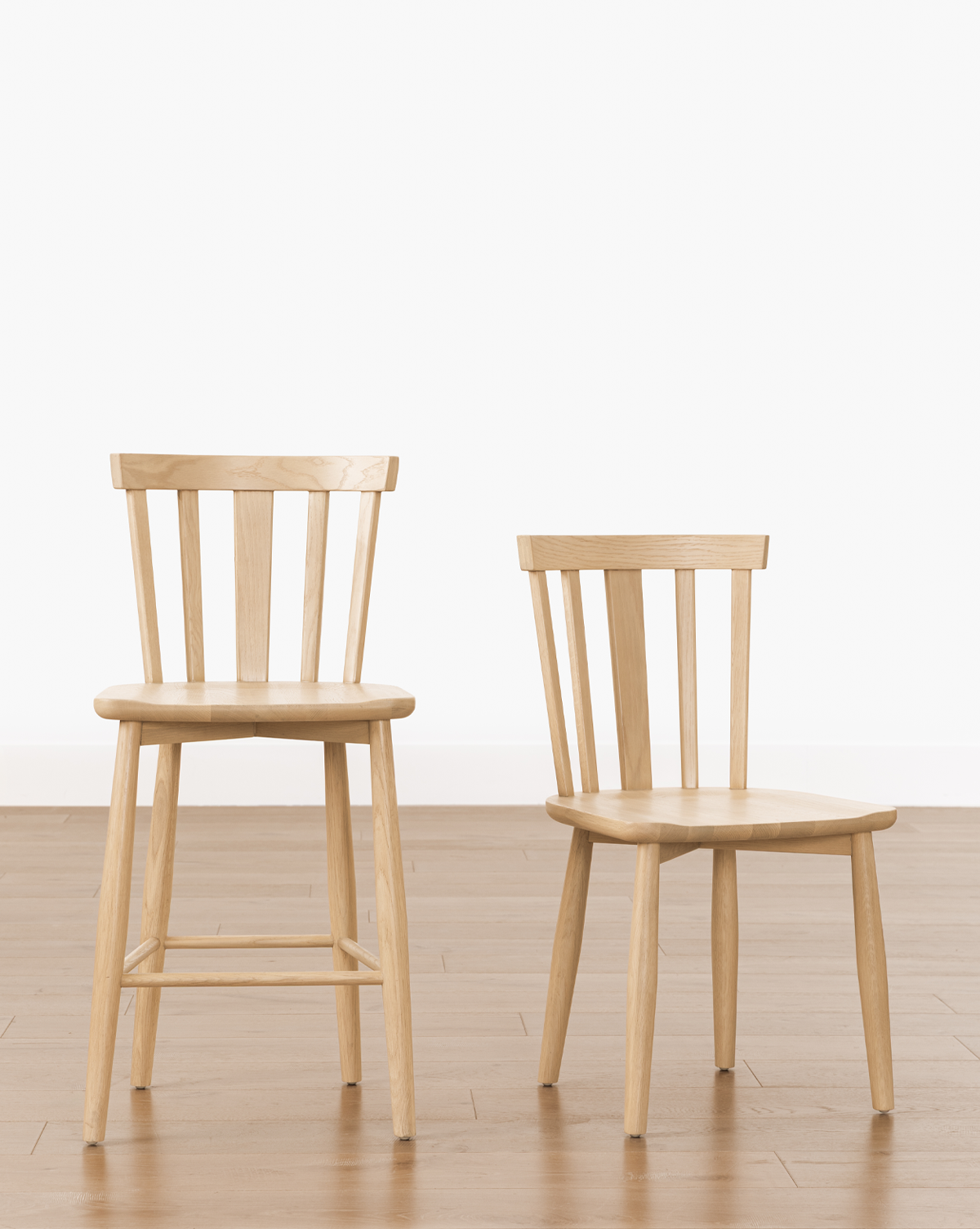 Two solid wood chairs, including a tall Hemingway Stool and a shorter chair, both with vertical slatted backrests, are side by side on a light wood floor against a plain white background, by McGee & Co.