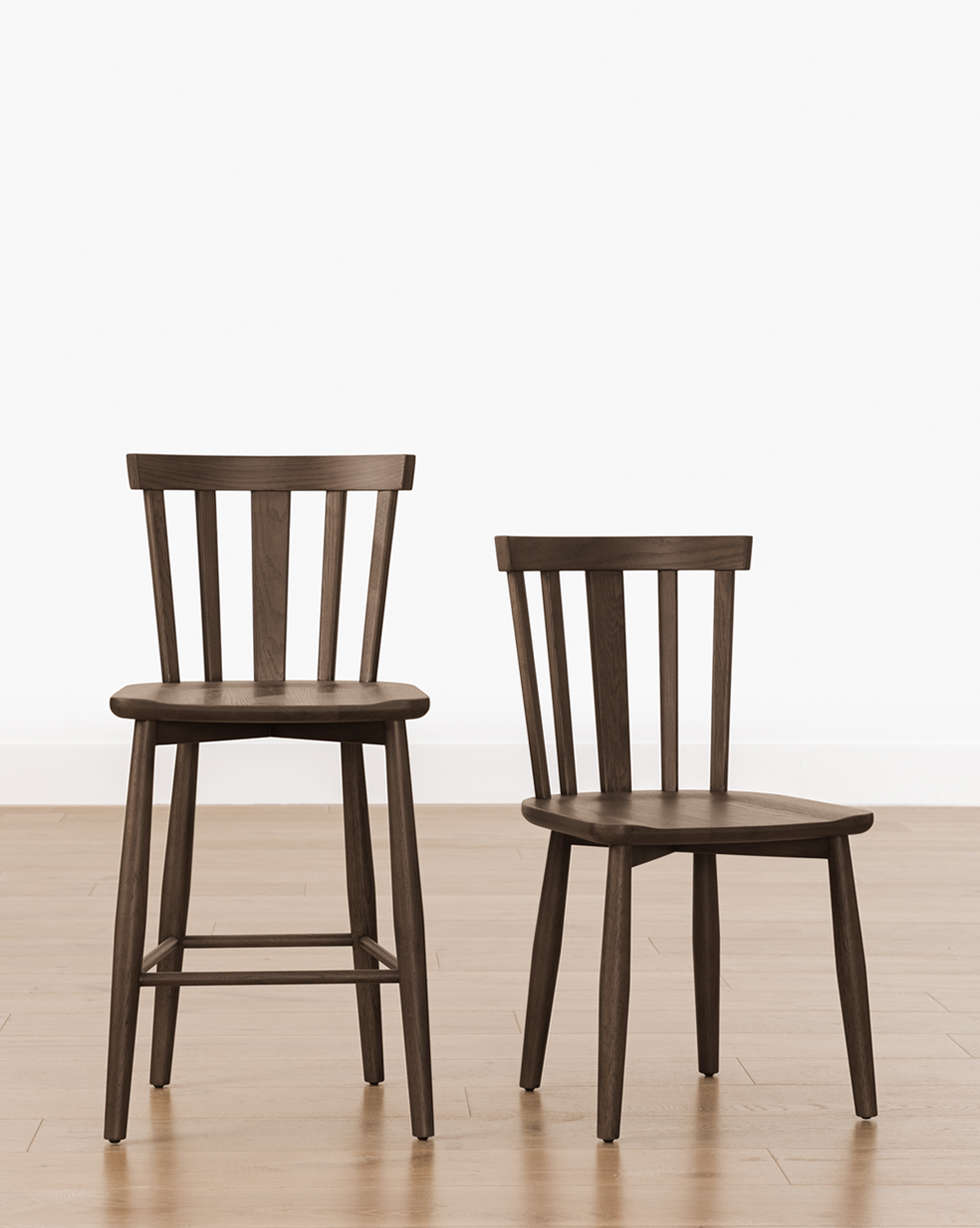 The Hemingway Stool, a dark solid wood chair with vertical slats on the backrest, stands tall for kitchen counters next to a standard-height chair. Both sit side by side on a light wooden floor against a plain white background, by McGee & Co.