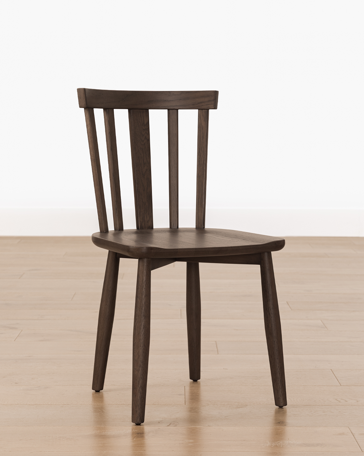 The Hemingway Chair is a versatile dining chair with a dark wooden finish, featuring a curved backrest with vertical slats. It's shown on a light wood floor against a plain white wall, by McGee & Co.