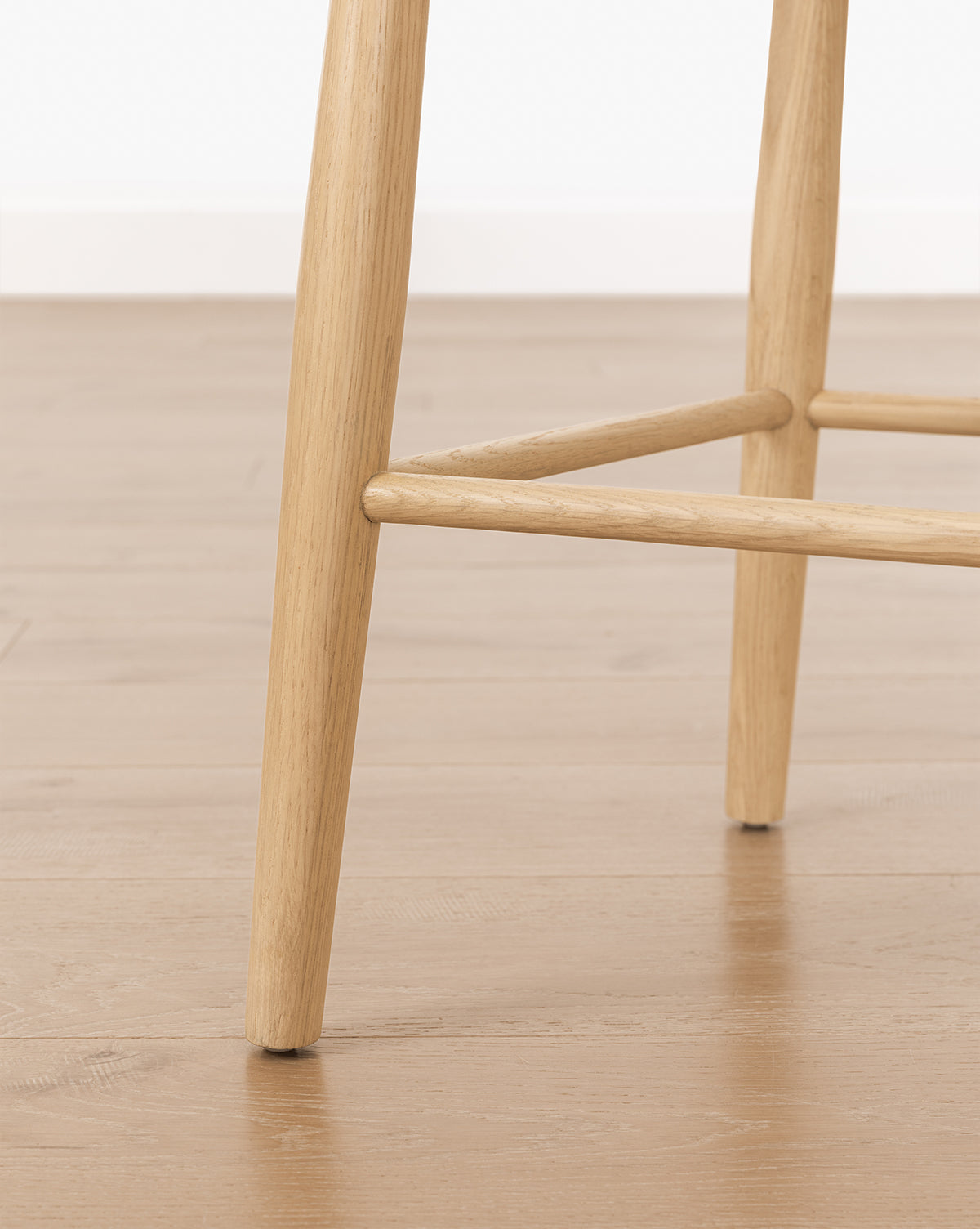 Close-up of the Hemingway Stool’s solid wood legs on a light brown wooden floor, with white baseboard and wall in the background, by McGee & Co.