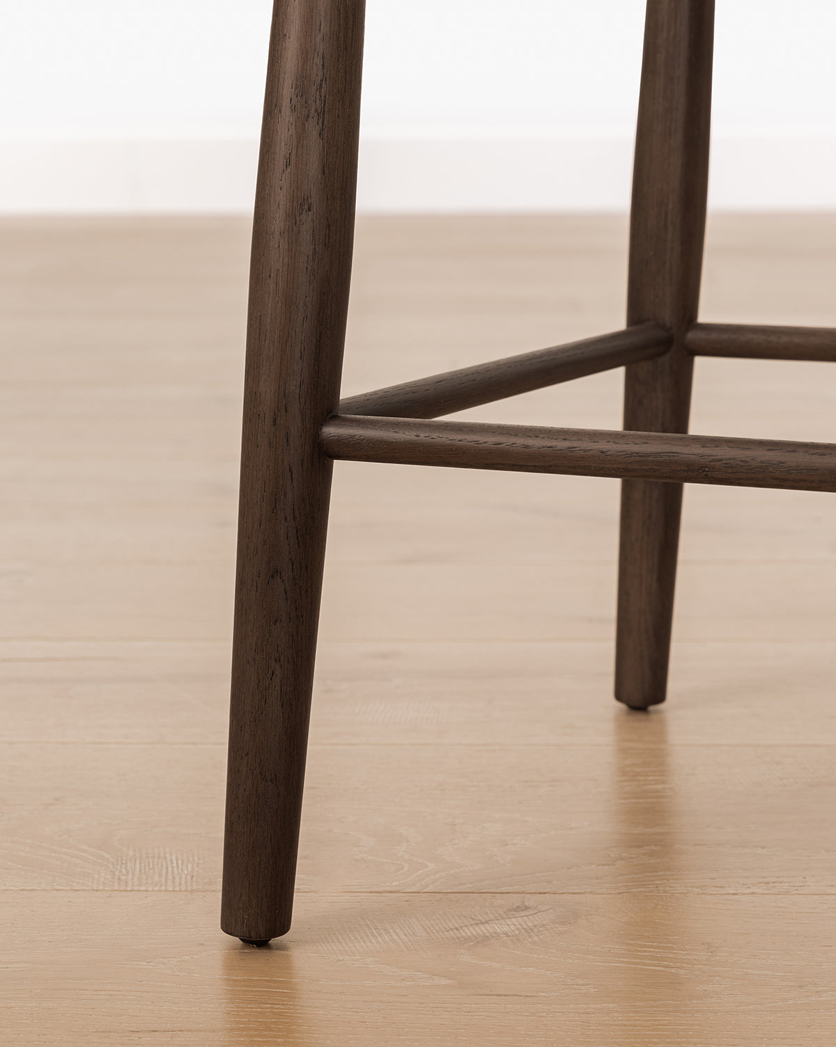 Close-up of the Hemingway Stool’s solid wood legs and crossbars on a light wood floor, ideal for kitchen counters with a white wall backdrop, by McGee & Co.
