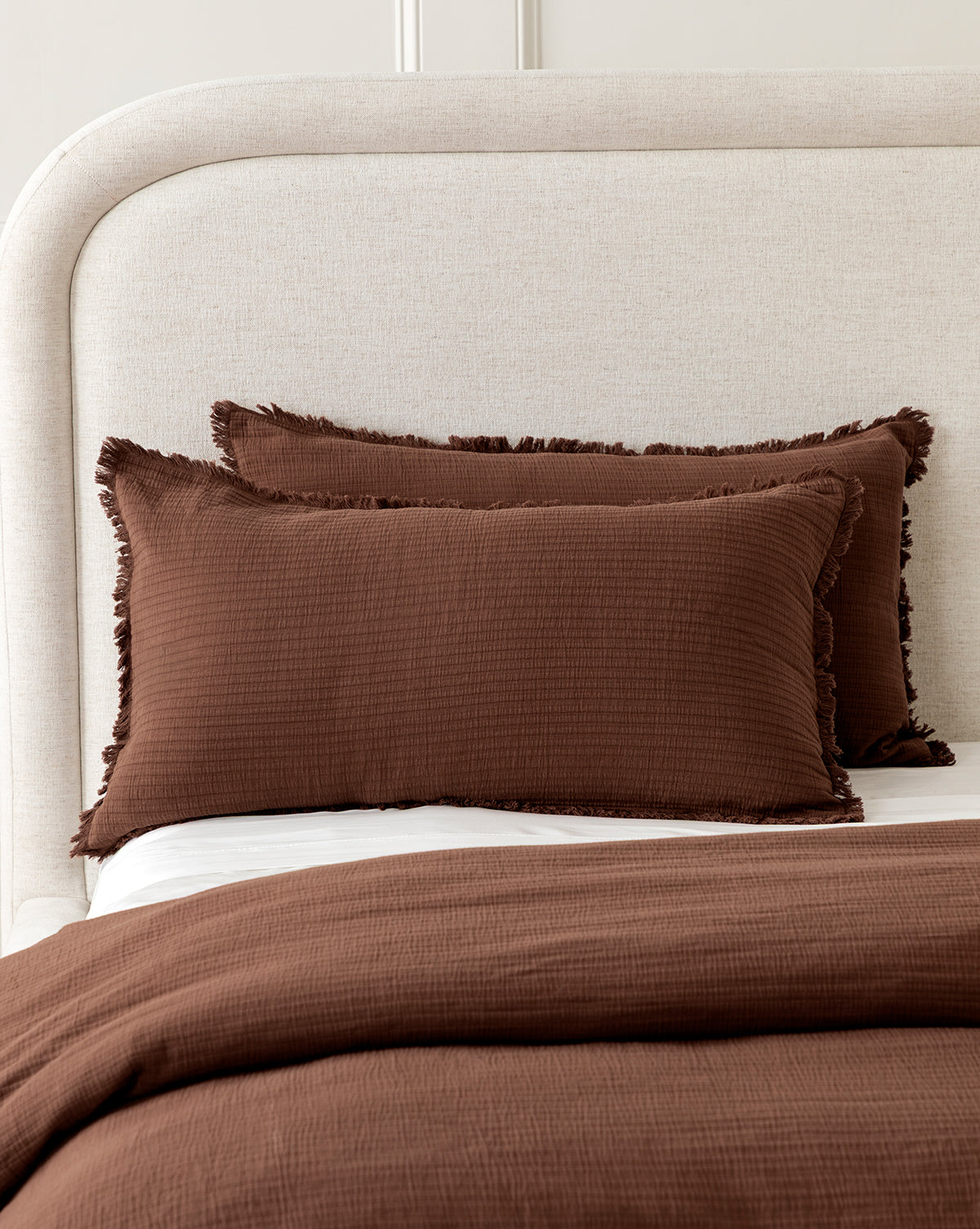 A neatly made bed with a light beige upholstered headboard, featuring Hendricks Chocolate Cotton Shams (Set of 2) with fringe edges and a matching brown textured cotton blanket, creates an inviting bedding ensemble.