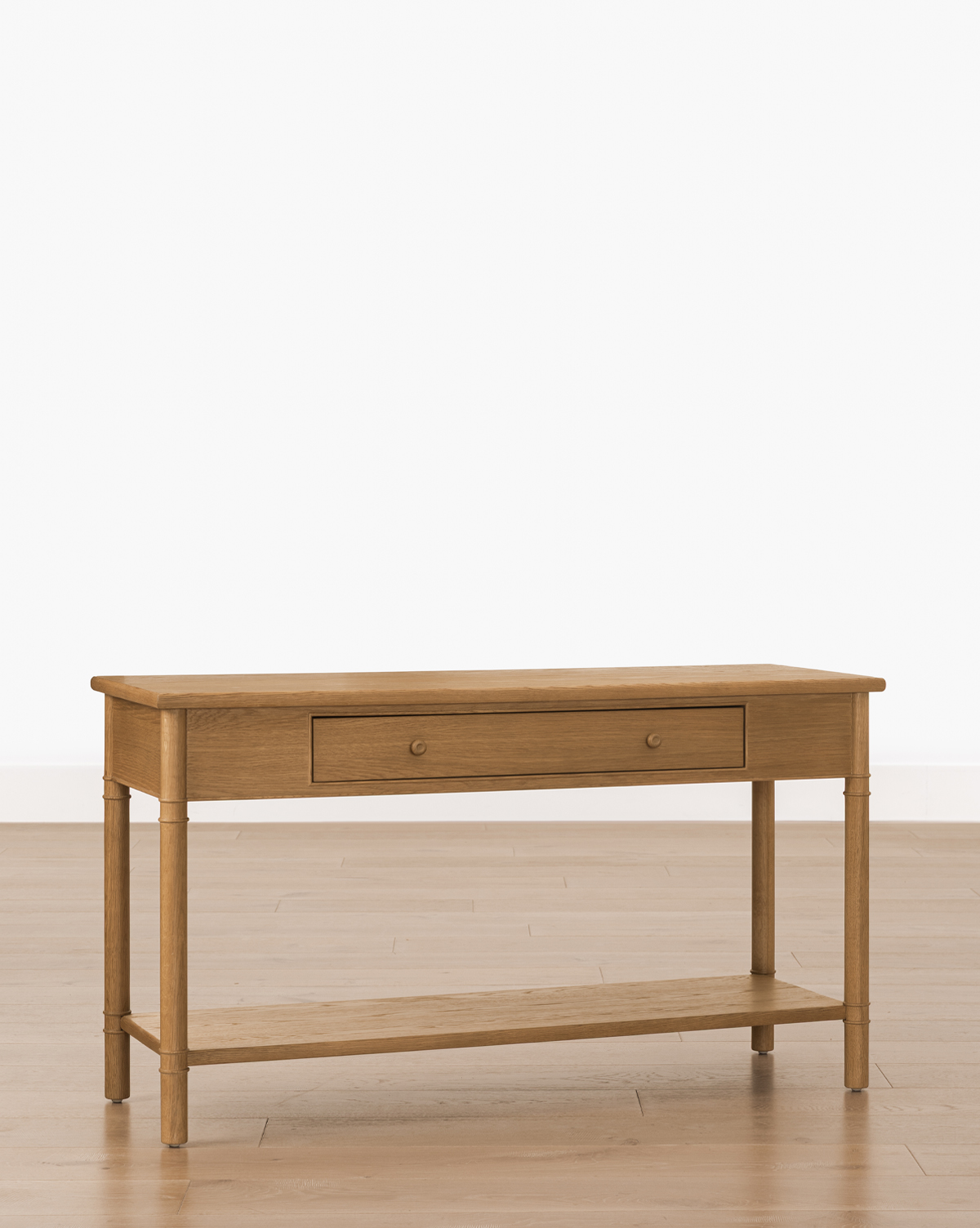 The Hindley Console features an oak finish, single central drawer, four legs, and a lower shelf for stylish storage. It is displayed on a light wooden floor against a plain white background, by McGee & Co.