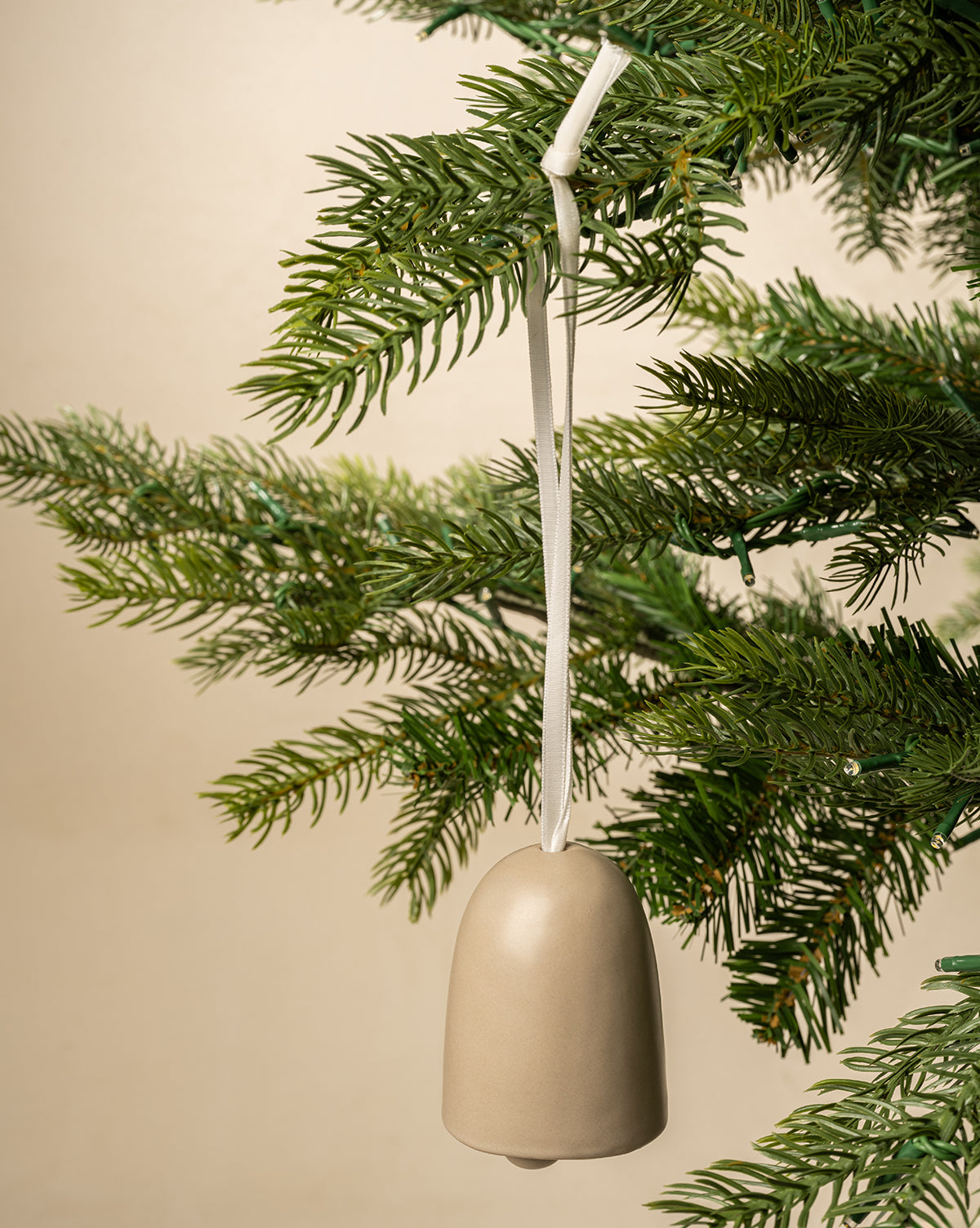 The Ivory Ceramic Bell hangs from a white ribbon on a green pine branch, bringing classic charm to your holiday decor with its simple design and elegant ivory finish, by McGee & Co.