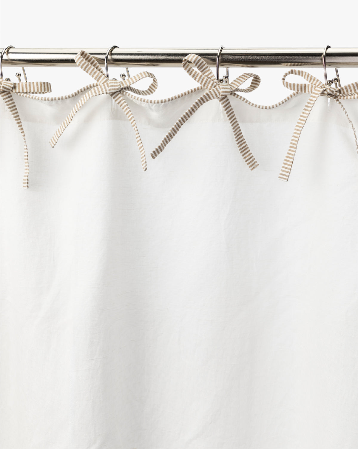 The Lillian Shower Curtain with Striped Ties is a white linen curtain that hangs from a metal rod, secured at the top with decorative beige and white striped ribbon ties fashioned into bows, by McGee & Co.