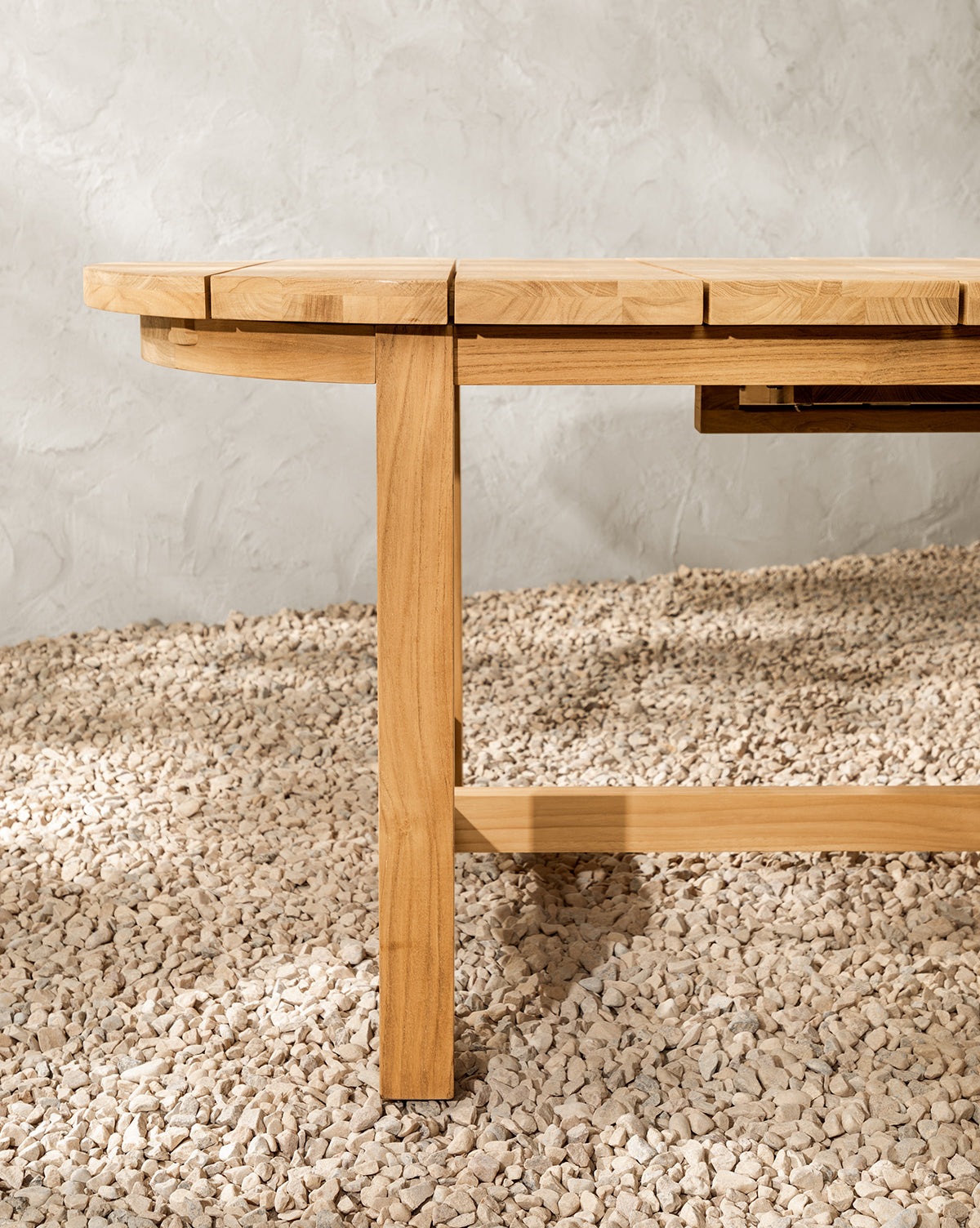 A close-up of the Linwood Teak Extension Dining Table leg and edge, crafted from light wood, shown on a textured, light-colored pebble floor with a gray textured wall in the background, by McGee & Co.