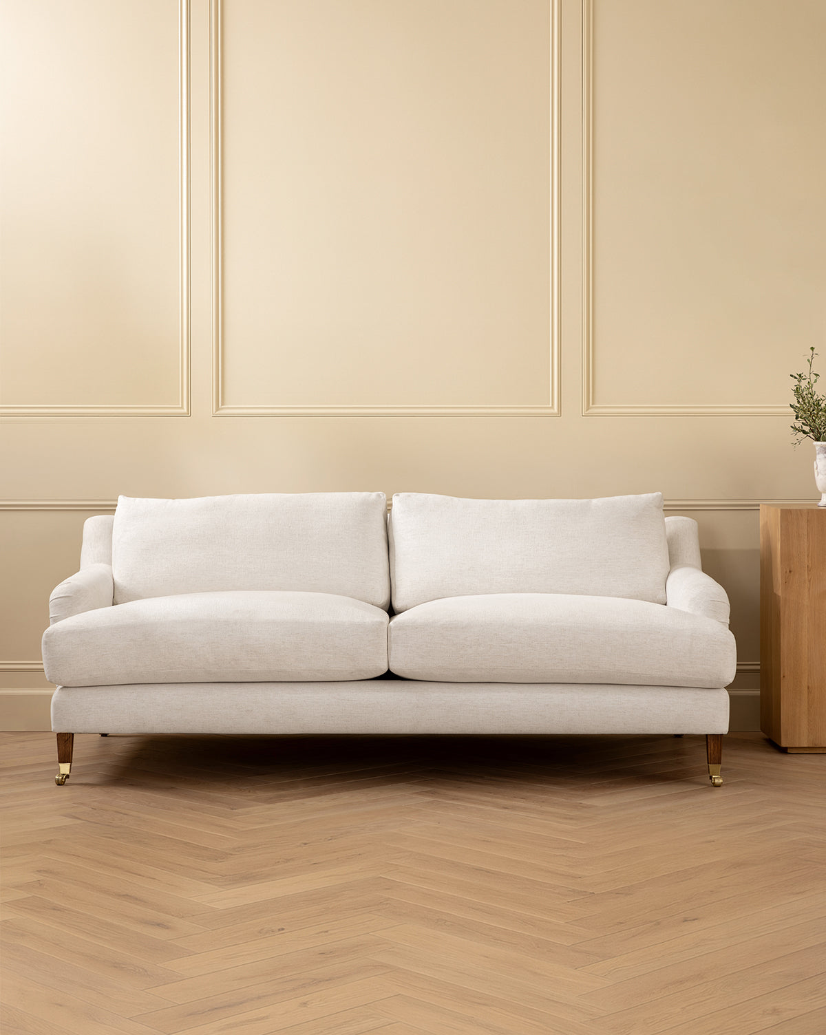 The Lucille English Roll Arm Sofa 90.5 in a modern cream hue, with plush down-wrapped cushions, sits before a beige paneled wall on light herringbone wood flooring. To the right, a small wooden side table holds a vase of greenery by McGee & Co.