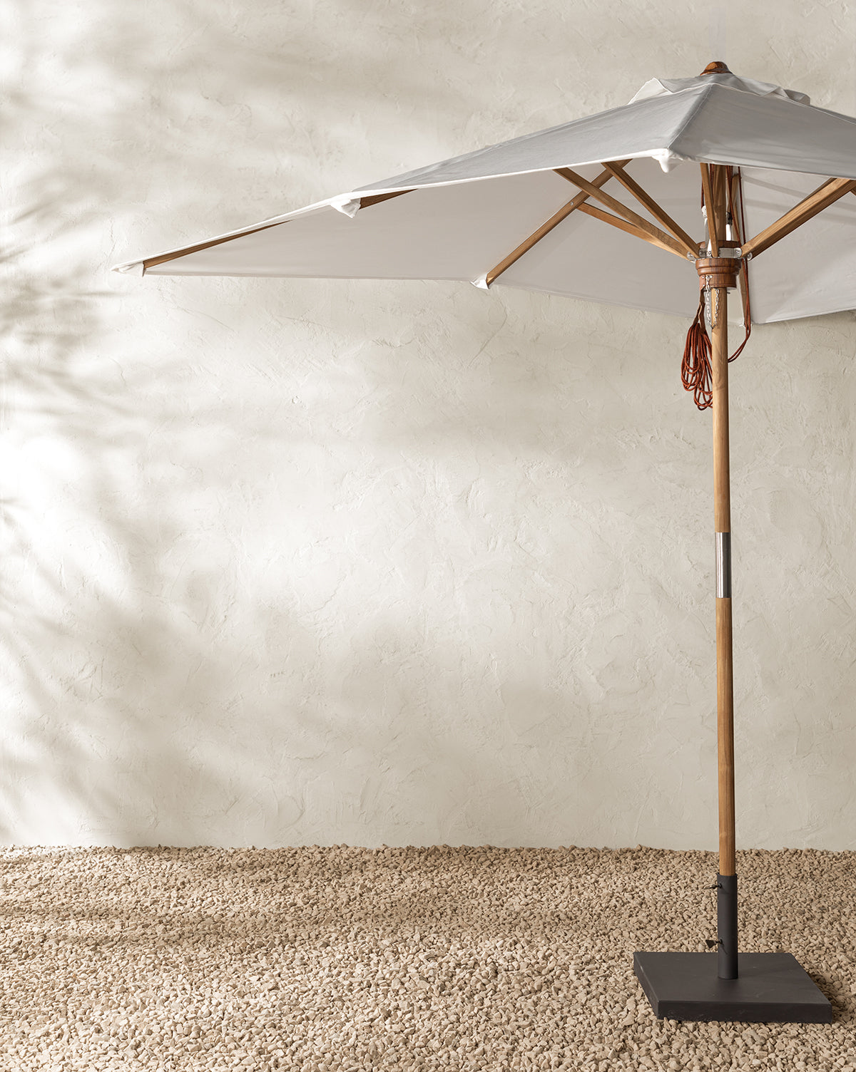 The Marques Outdoor Rectangular Umbrella, featuring a large white canopy and teak pole, stands on a textured gravel surface against a light beige stucco wall, casting soft shadows in this minimalist outdoor space, by McGee & Co.