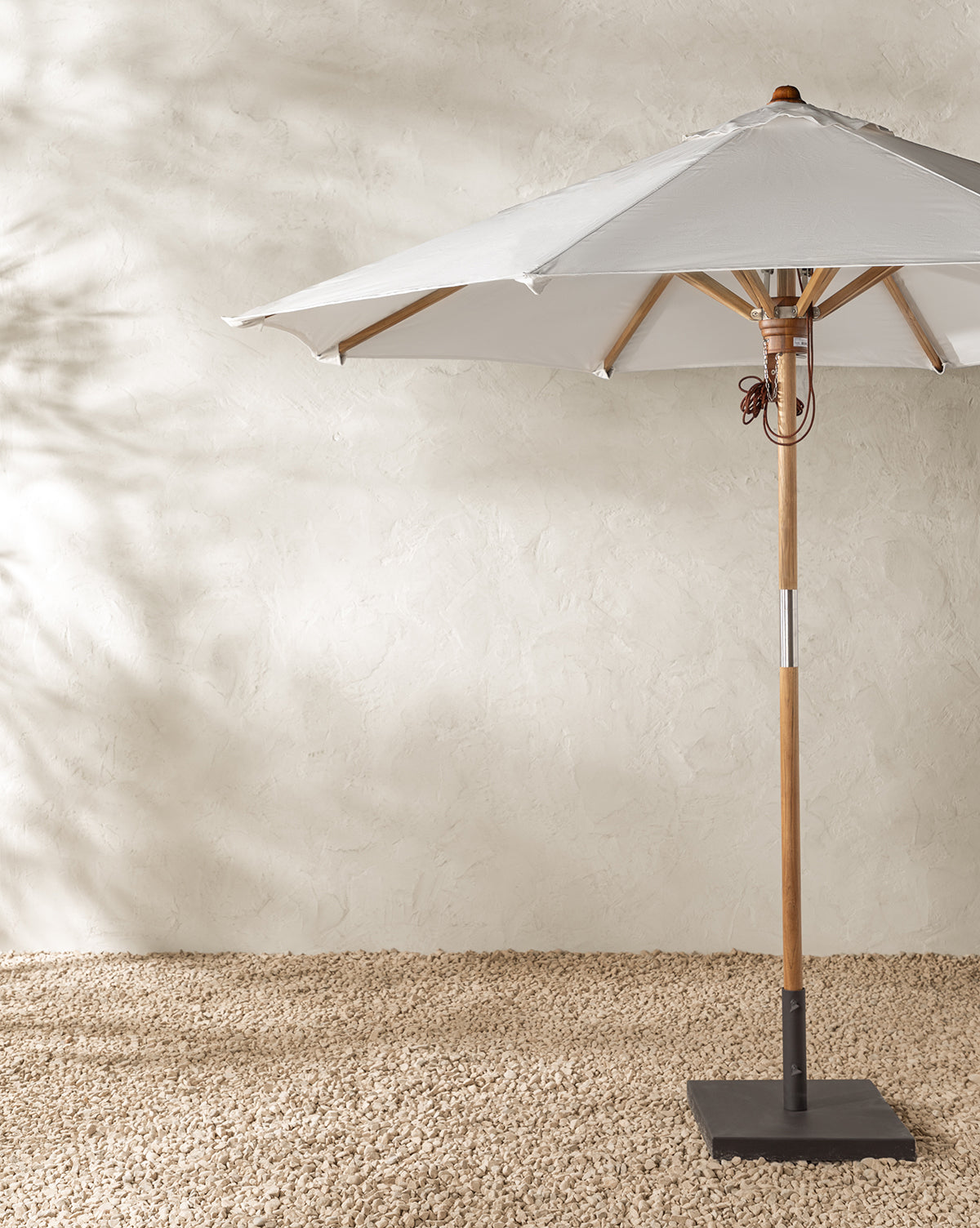The Marques Outdoor Round Umbrella features a weather-resistant acrylic white canopy and sturdy teak pole, standing on textured gravel against a light beige stucco wall with soft background shadows, by McGee & Co.