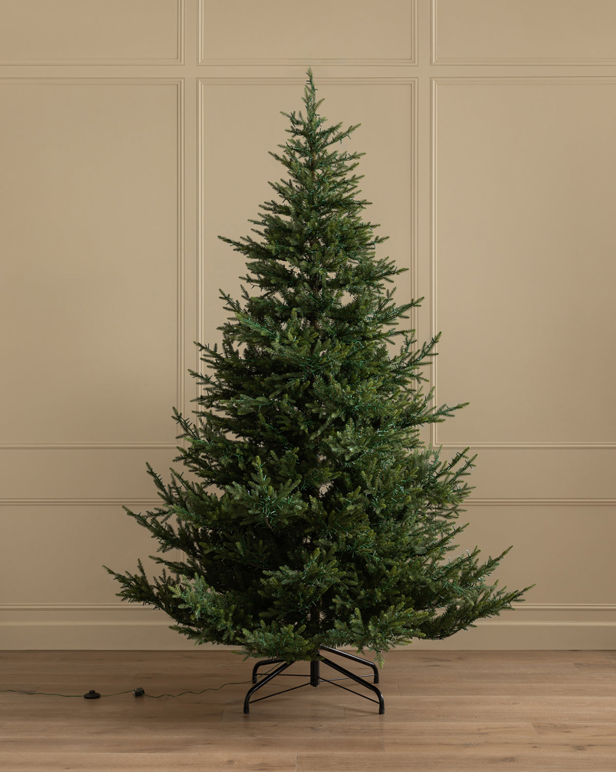 A 9' Norway Spruce Pre-Lit Faux Tree with dense green branches stands on a metal stand in a plain room with beige paneled walls and wooden floor, an electric cord lying nearby, by McGee & Co.