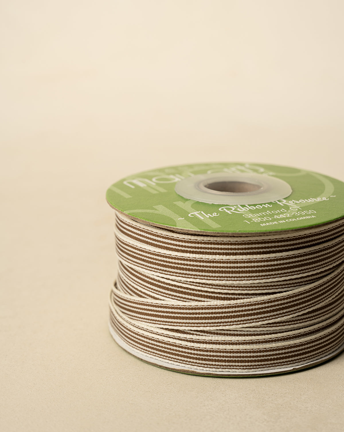A spool of 3/8" Oatmeal & Brown Woven Grosgrain Stripe Ribbon, neatly wound with a green label on top, rests on a beige surface—ideal for holiday decor or gift wrapping, by McGee & Co.