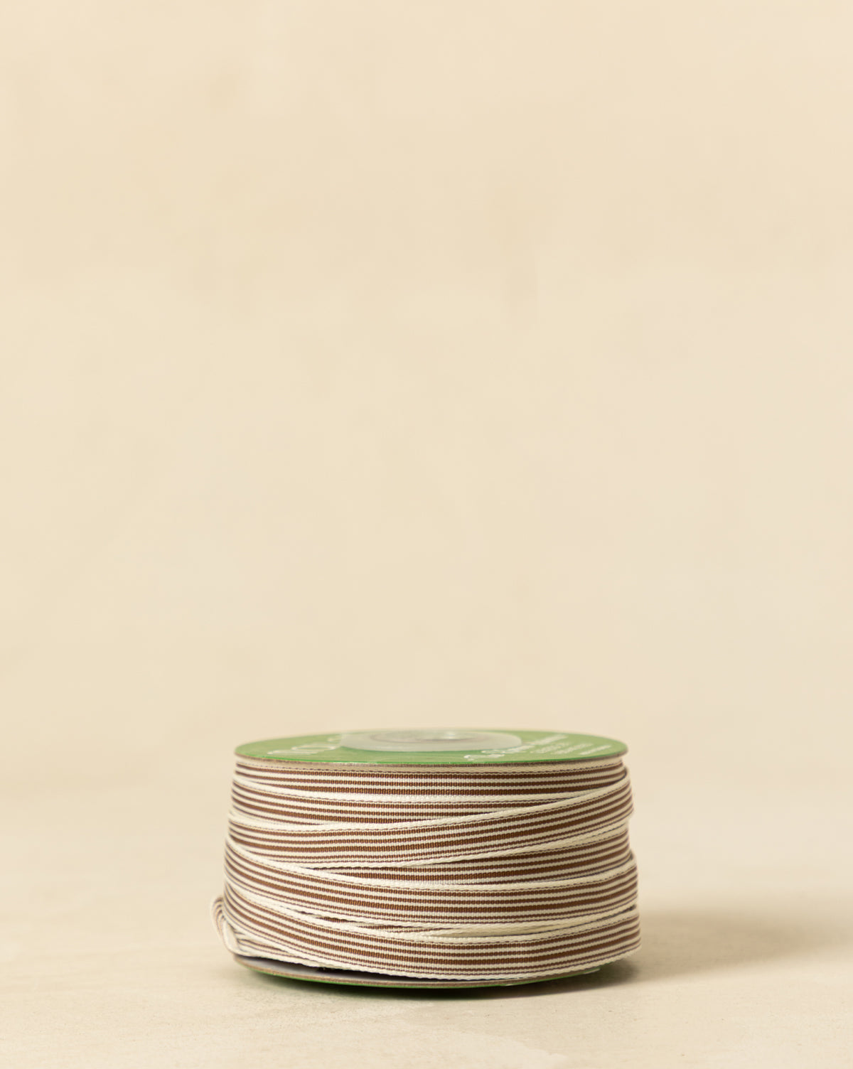 A spool of 3/8" Oatmeal & Brown Woven Grosgrain Stripe Ribbon rests on a light beige surface with a plain background—ideal for holiday decorating, by McGee & Co.