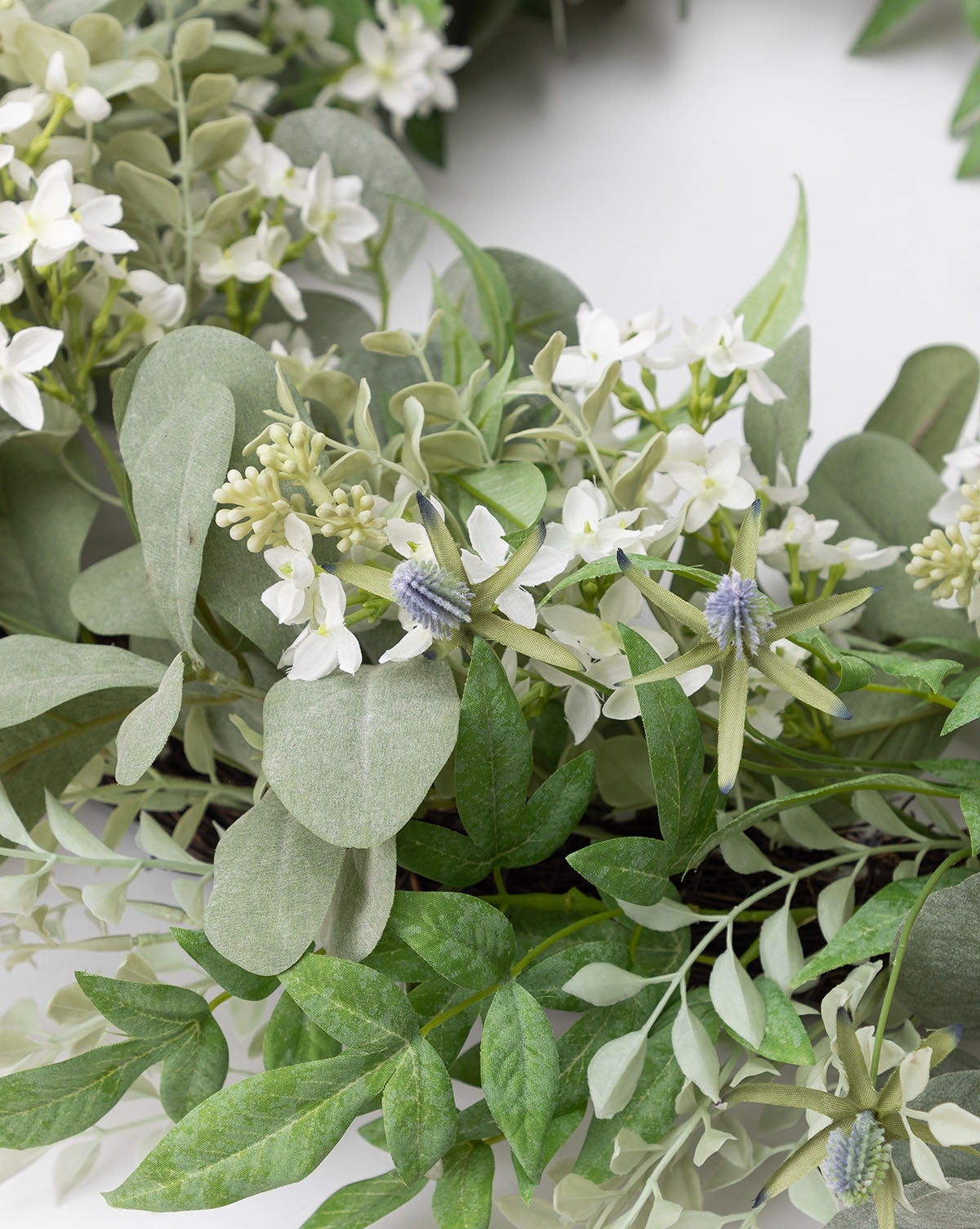 A close-up of the Orange Jasmine & Eucalyptus Wreath showcases green leaves, eucalyptus, small white flowers, and touches of light purple thistle against a white background.