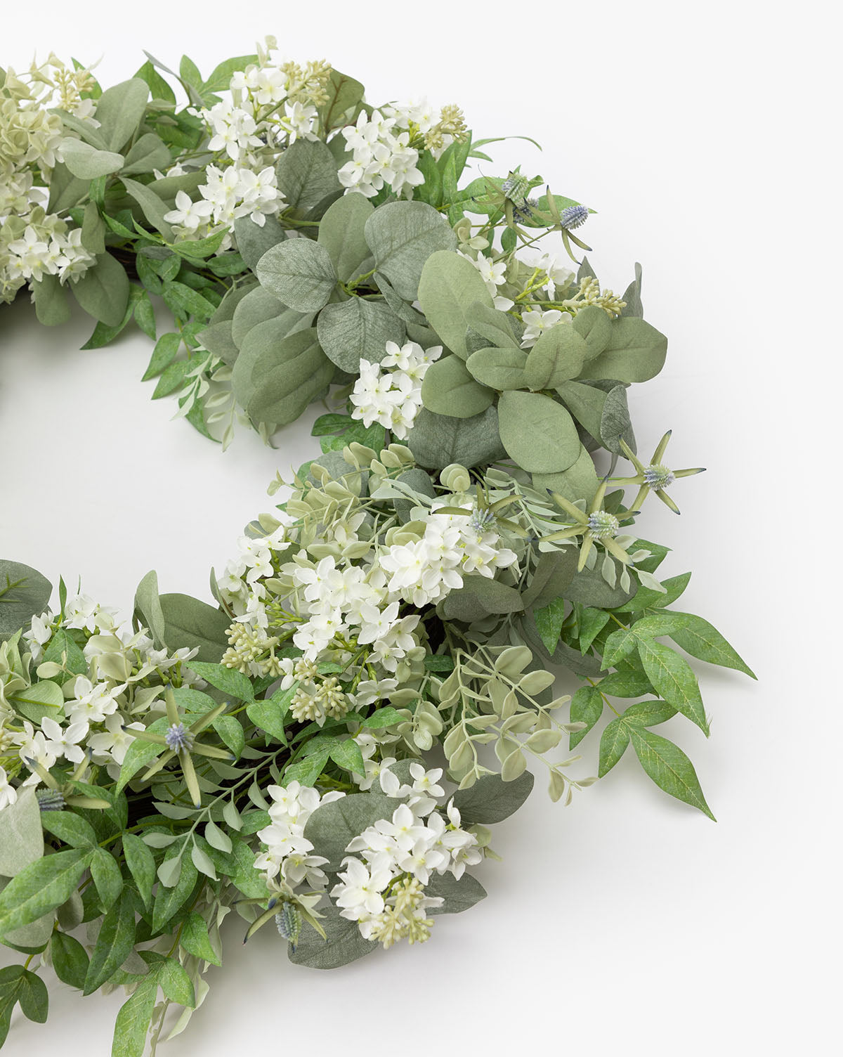 A close-up of the Orange Jasmine & Eucalyptus Wreath, featuring green leaves and small white flowers in a lush, natural style against a plain white background.