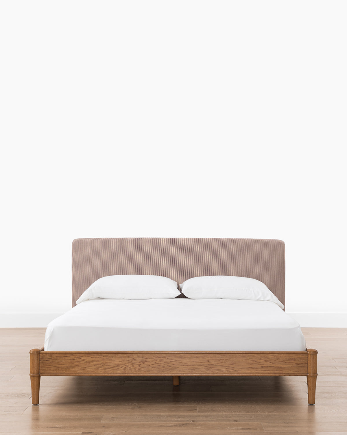 The Palladio Bed by Makerspalm features a wooden frame with honey oak legs and a light brown upholstered headboard, dressed in white sheets and pillows, set on a wooden floor against a plain white wall, finished with accents from McGee & Co..