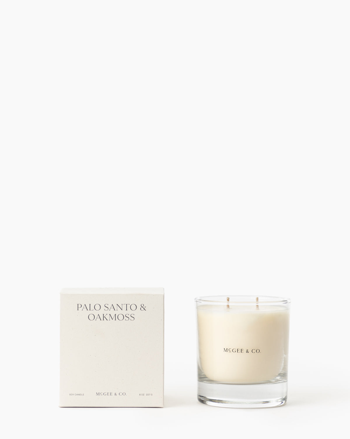 A white box labeled Palo Santo & Oakmoss Candle stands beside a glass luxury soy candle with two wicks, both displaying minimalist McGee & Co. branding on a white background, by McGee & Co.