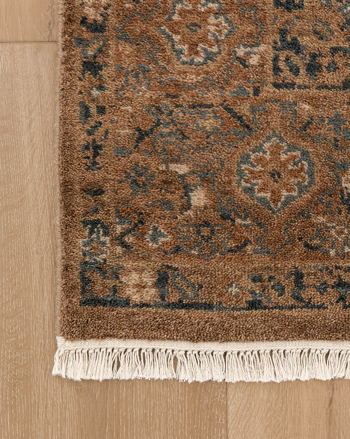 A close-up of the Piedmont Hand-Knotted Wool Rug, showing its brown and blue traditional motifs with white fringes, displayed on a light wooden floor, by McGee & Co.