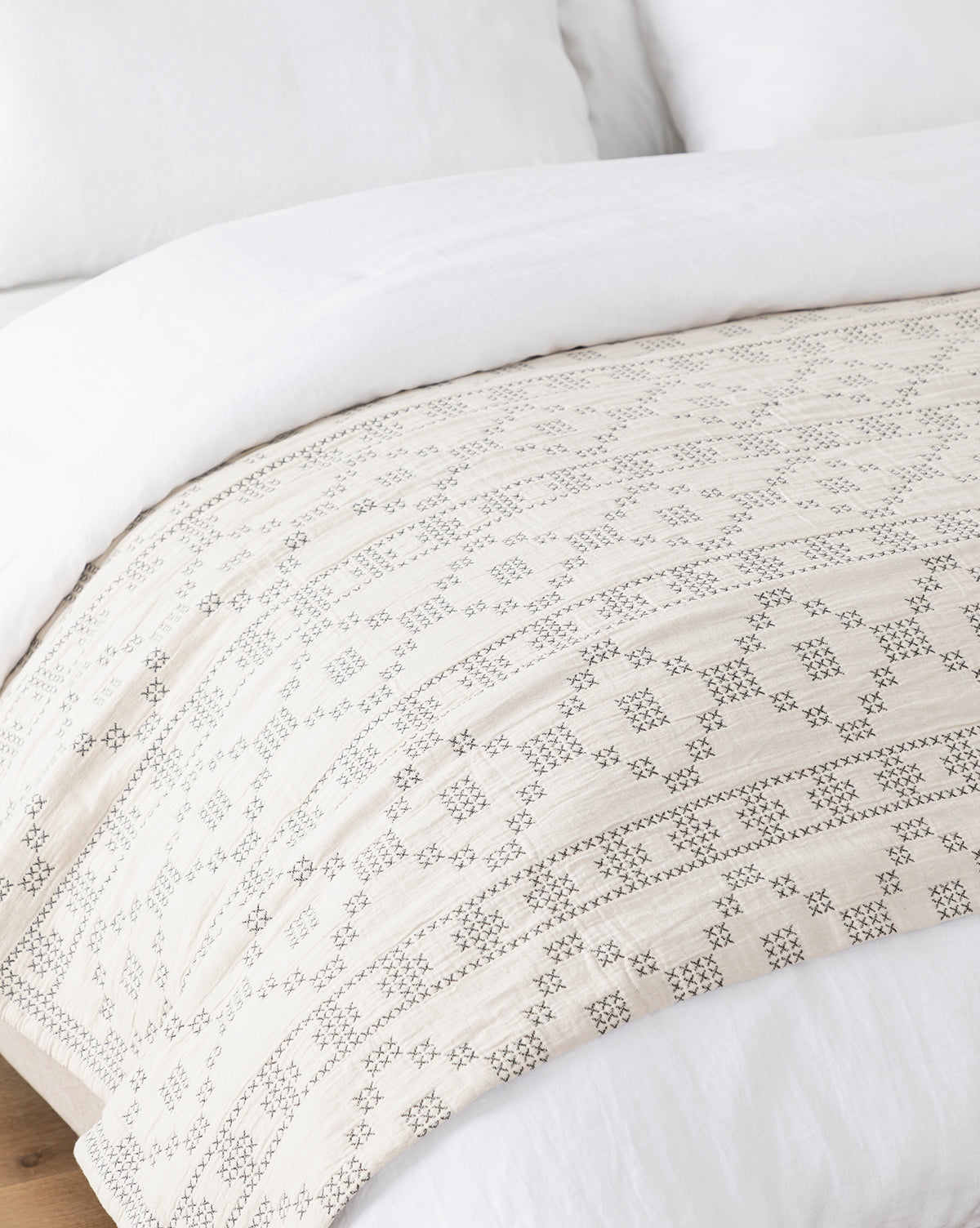 A close-up of a bed with crisp white bedding and the Prescott Coverlet, showcasing a geometric black embroidered pattern, draped across the foot of the bed on a wooden floor, by McGee & Co.