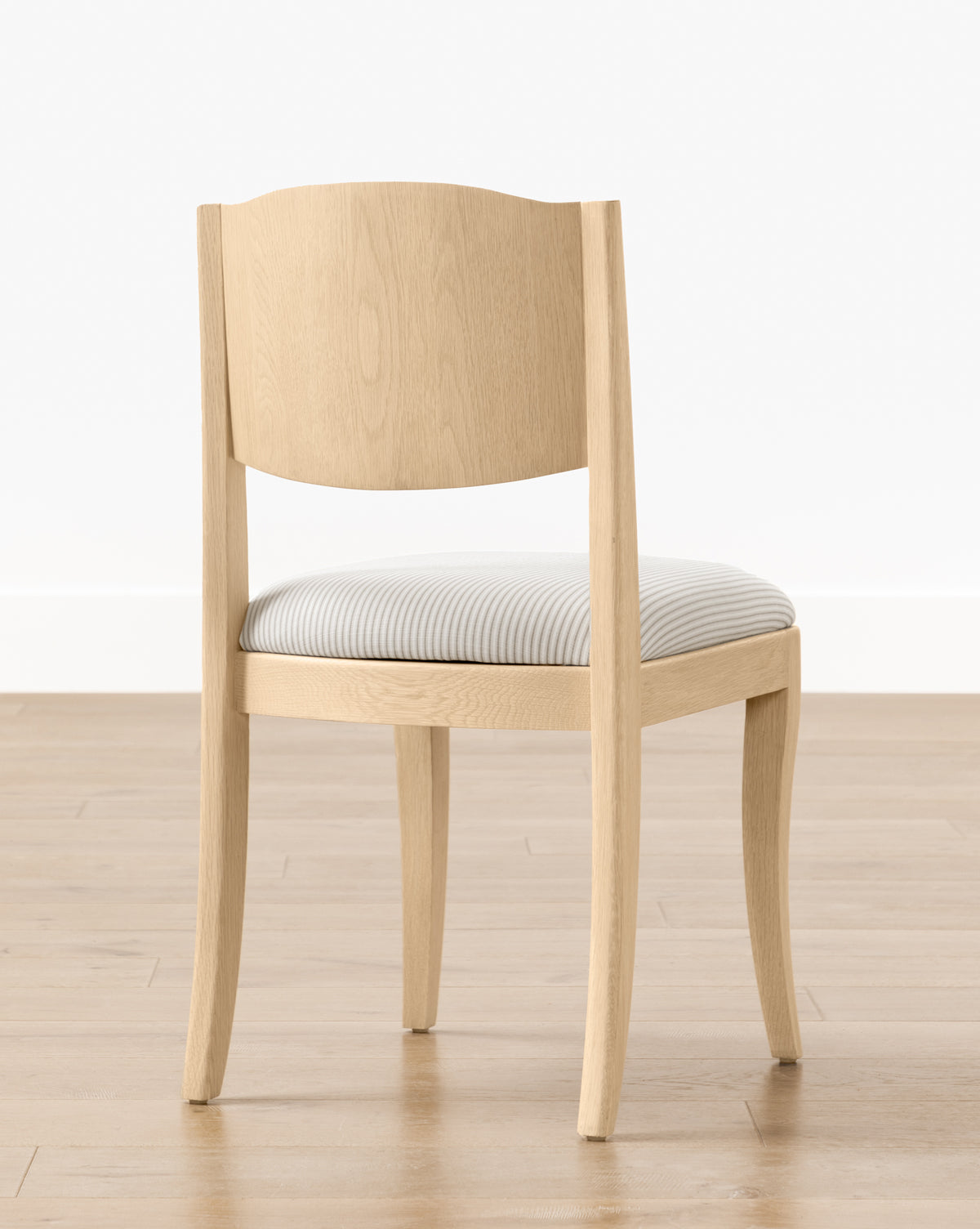 The Shepherd Dining Chair features light wood, a curved backrest, four legs, and a white and gray striped upholstered seat cushion. It is shown on a light wooden floor with a white wall background by McGee & Co.