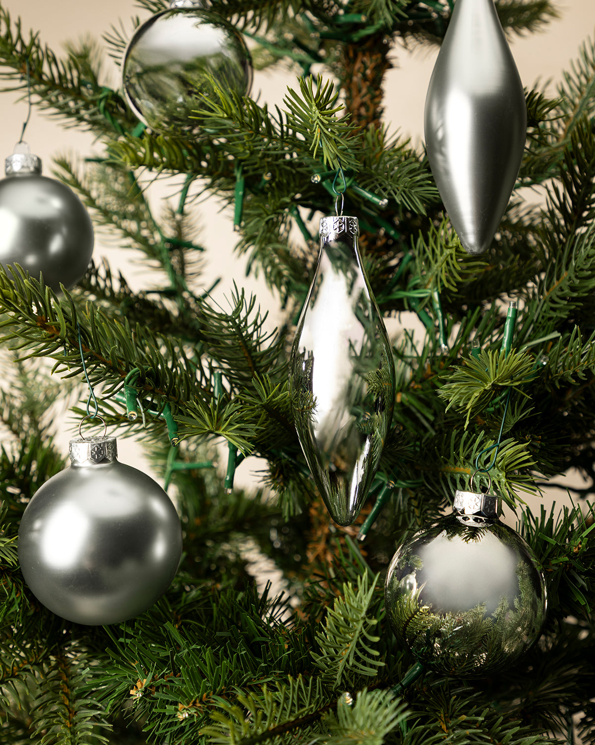 A close-up of a holiday tree adorned with Assorted Ornaments Silver (Set of 10), featuring shiny round and teardrop-shaped designs nestled among green pine needles against a neutral background, by McGee & Co.