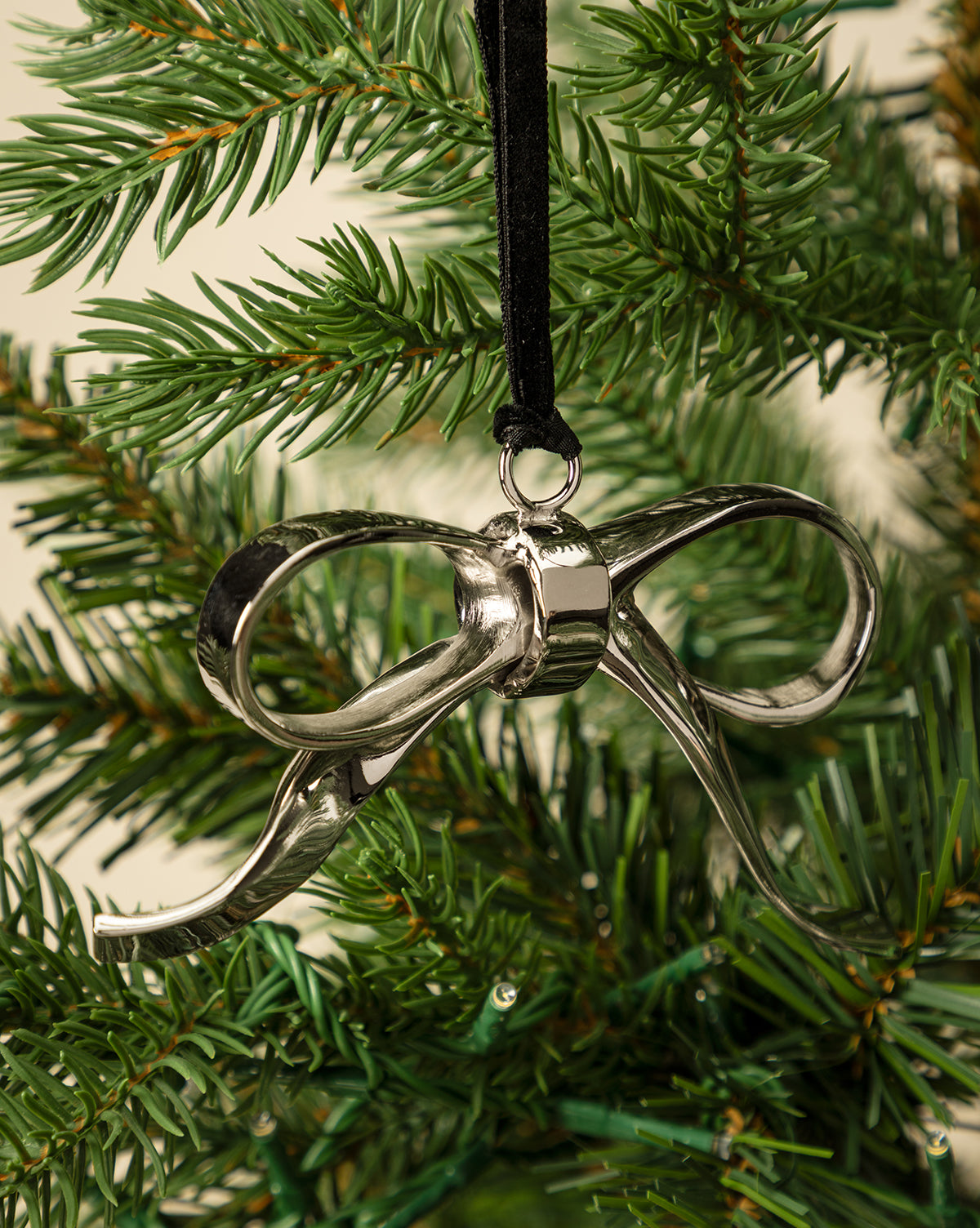 The Silver Bow Ornament, suspended from a black ribbon on a green holiday tree, brings festive charm and elegant décor to your seasonal celebrations, by McGee & Co.