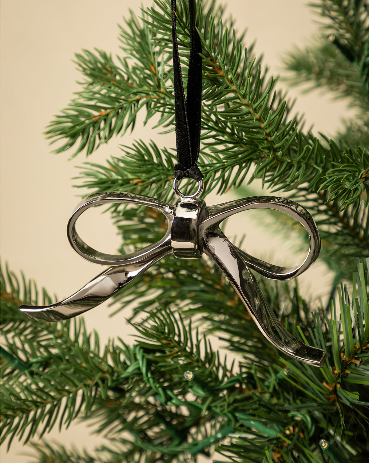 The Silver Bow Ornament hangs from a black ribbon on a green holiday tree, creating festive décor against a plain beige background, by McGee & Co.