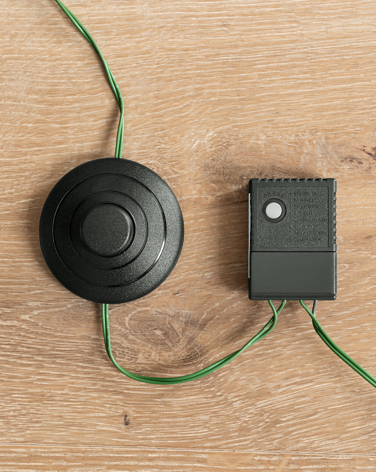 A round black wired foot pedal switch and a small rectangular control box with green wires from the 7' Snowy Grandis Fir Pre-Lit Faux Tree are both placed on a light wood surface, by McGee & Co.