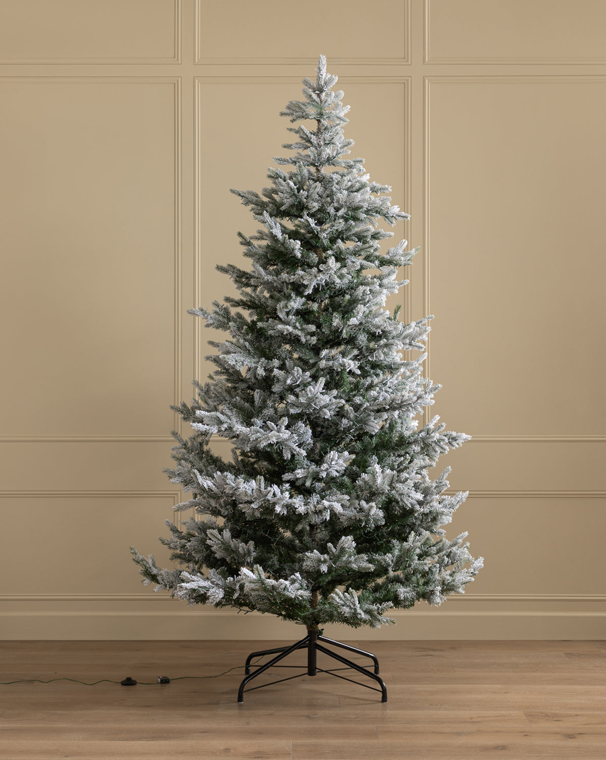 A 9' Snowy Grandis Fir Pre-Lit Faux Tree stands on a black metal stand in a room with beige paneled walls and a wooden floor, with an electrical cord visible near the tree’s base, by McGee & Co.