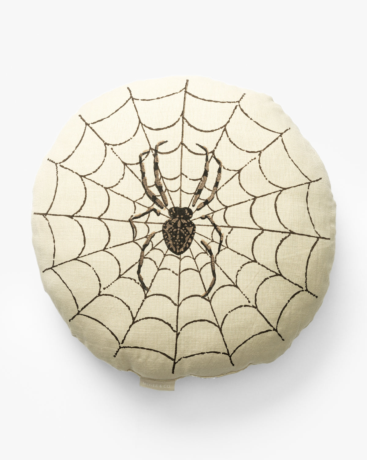 The Spider Pillow is a round beige cushion with a black spider and an embroidered web design covering the surface—an ideal Halloween decor accent, by McGee & Co.