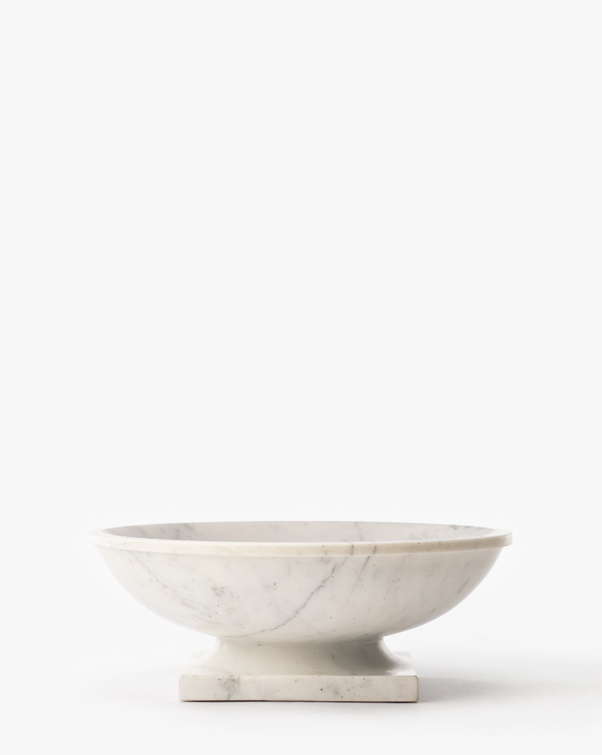 The Tayva Bowl is a sculptural white marble piece with subtle gray veining, featuring a shallow round form, wide rim, and square pedestal base, displayed on a plain white background by McGee & Co.