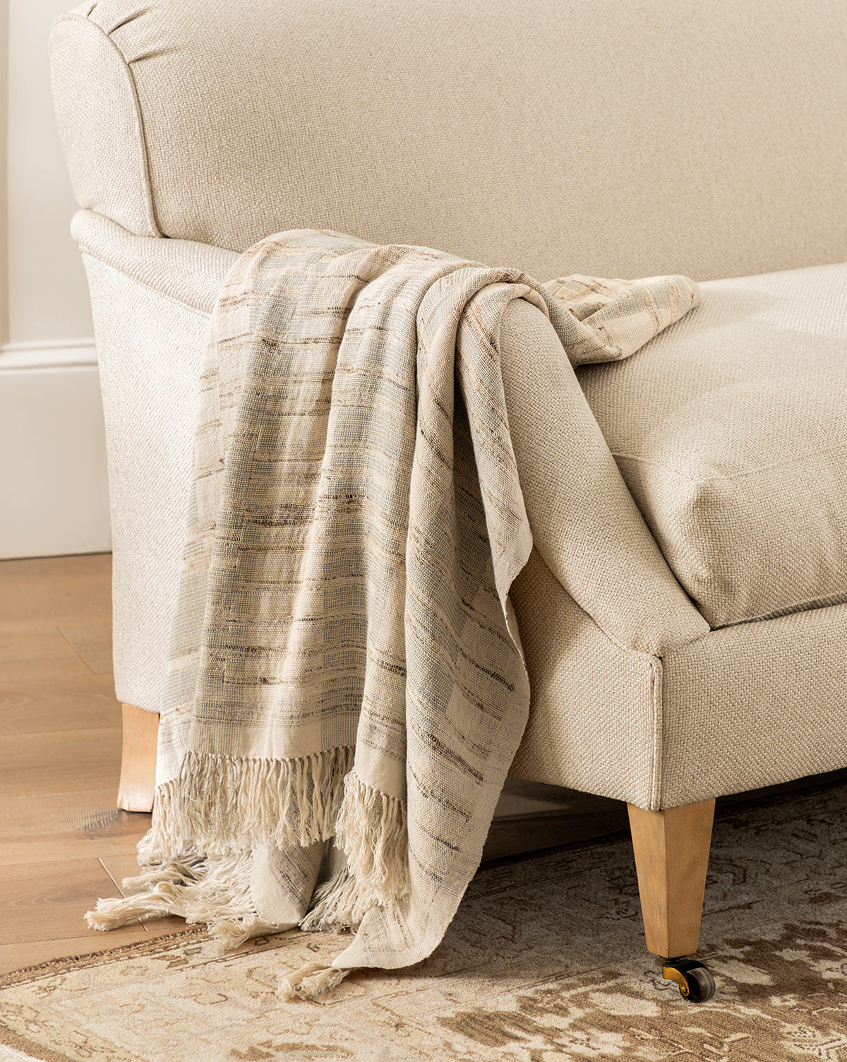 The Theodora Block Stripe Throw, made from beige cotton and raw silk with fringed edges, is draped over a light-colored upholstered sofa on a wooden floor beside a patterned rug.