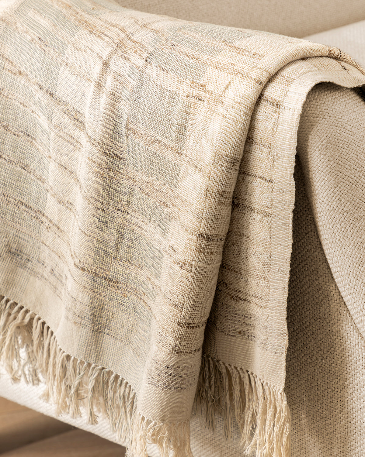 The Theodora Block Stripe Throw, a beige and cream textured blanket with tassels and a subtle plaid pattern, drapes over the arm of a light sofa to add a cozy accent to your furniture.