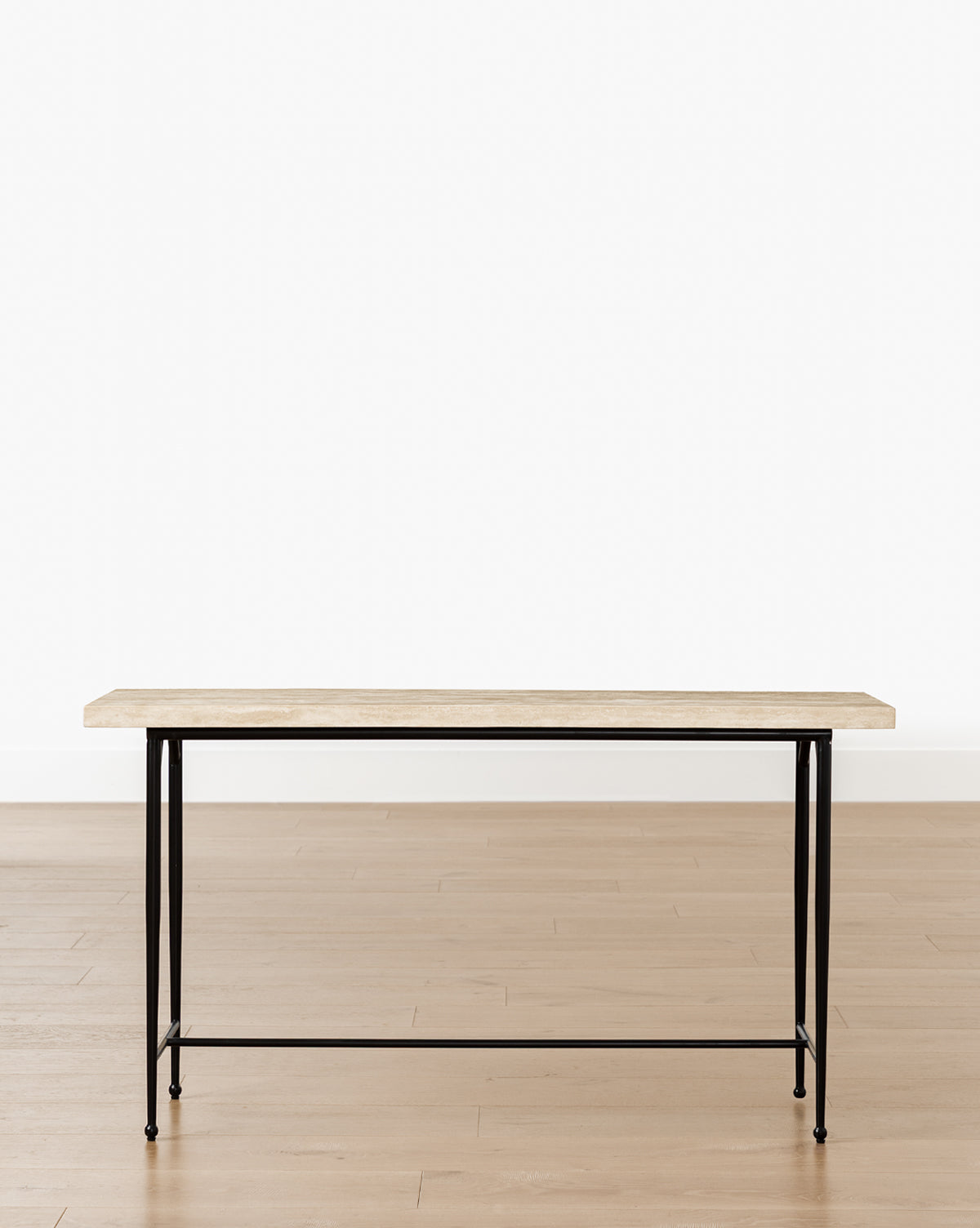 The Travertine Console features a minimalist design with a light wood top and slender black hand-forged iron legs, displayed on a light wood floor against a plain white background by McGee & Co.