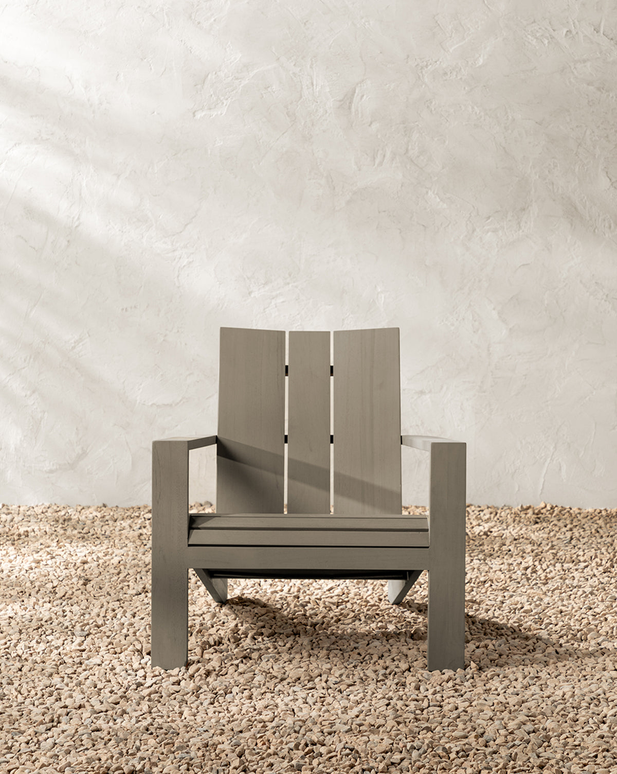 The Triby Gray Teak Outdoor Lounge Chair features wide armrests and a slatted backrest, placed on light pebbles against a textured beige wall—ideal for enhancing any patio space by McGee & Co.