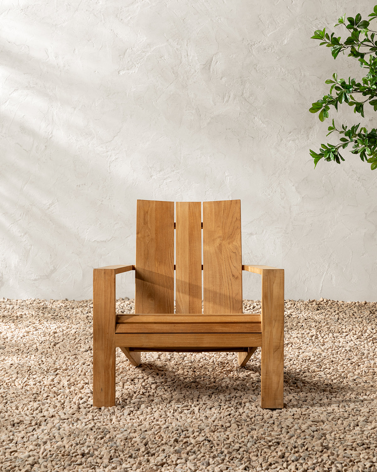 The Triby Teak Outdoor Lounge Chair rests on pale pebbles by a textured white wall, while sunlight creates gentle shadows and a leafy green plant peeks in from the right, highlighting this tranquil, all-weather setting, by McGee & Co.