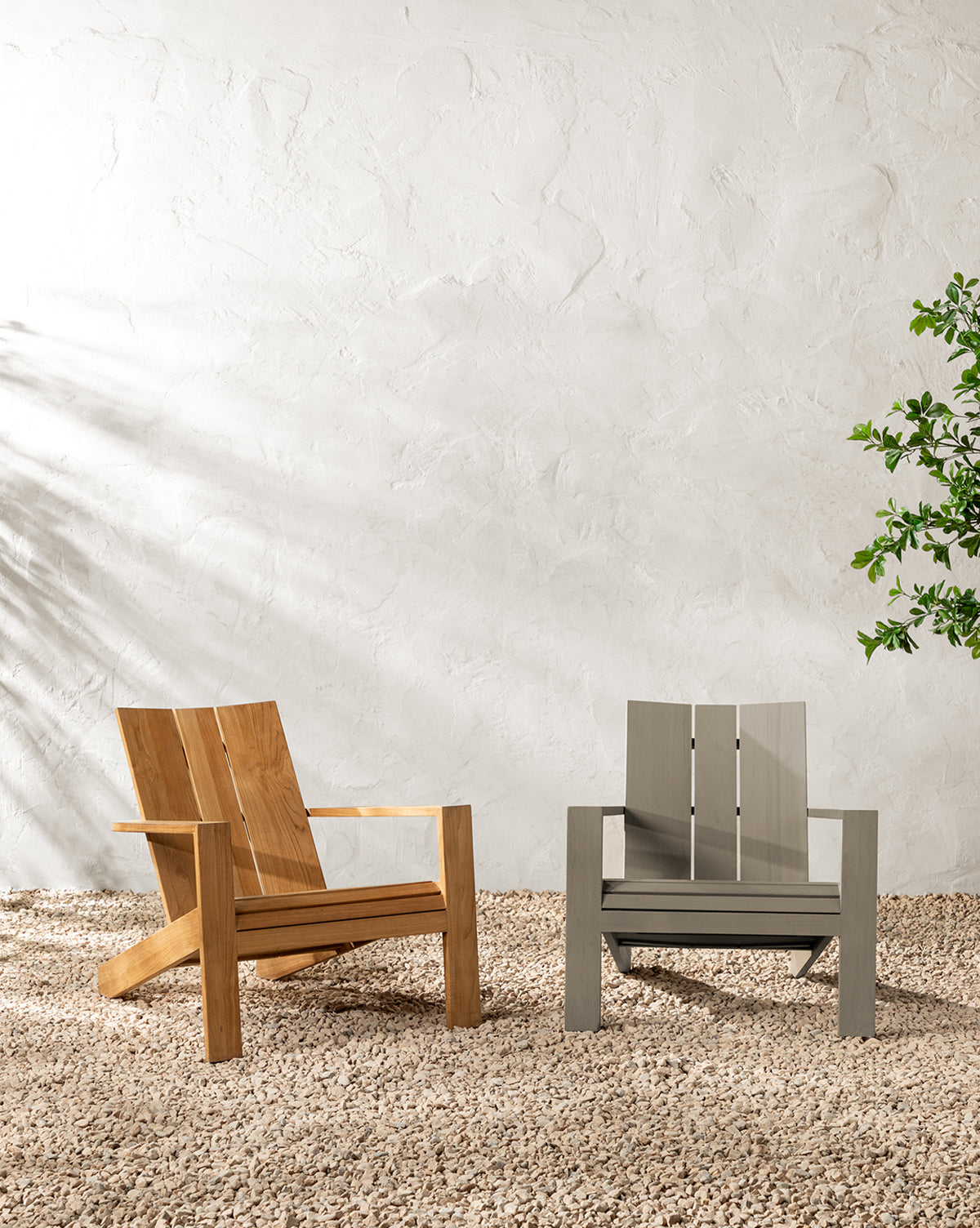 Two modern patio chairs, including the Triby Teak Outdoor Lounge Chair and a gray all-weather chair, sit on gravel against a textured white wall. A leafy plant to the right and sunlight create gentle shadows, enhancing the inviting scene by McGee & Co.