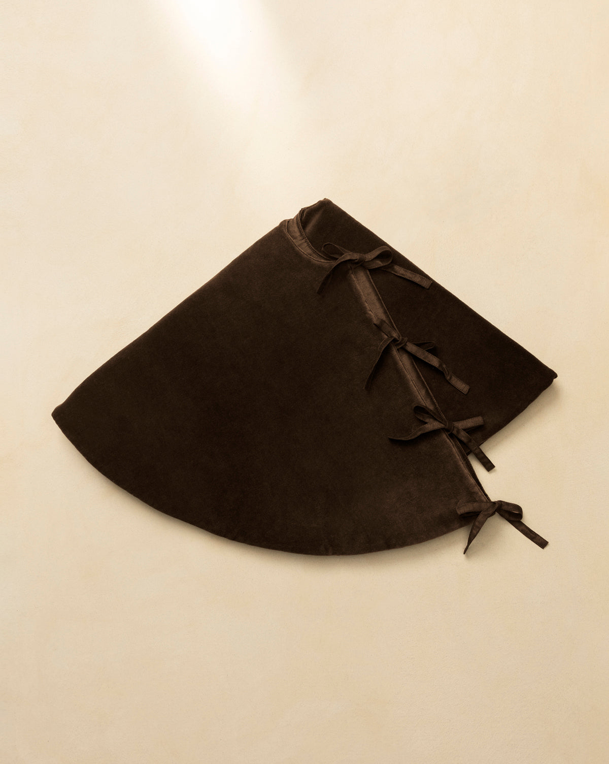 The Velvet Brown Tree Skirt, a dark brown semi-circular design with three ribbon tie closures, is displayed flat on a beige background, highlighting its luxurious texture—ideal for stylish Christmas tree decor, by McGee & Co.