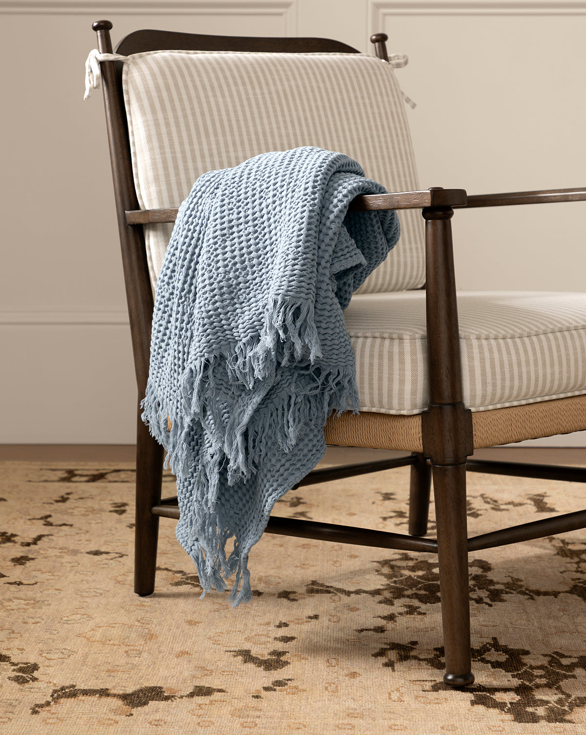 A striped upholstered chair with dark wood arms sits on a patterned beige rug; the Viola Throw, featuring a textured weave and light blue fringe, is draped casually over the chair’s arm and seat, by McGee & Co.