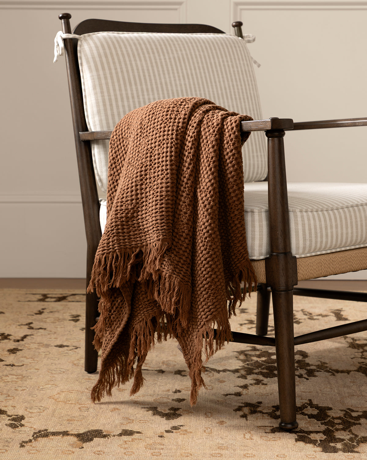 The Viola Throw, featuring a textured weave and fringe, is draped over a cushioned wooden chair with striped upholstery, positioned on a patterned beige rug in a neutral-toned room, by McGee & Co.