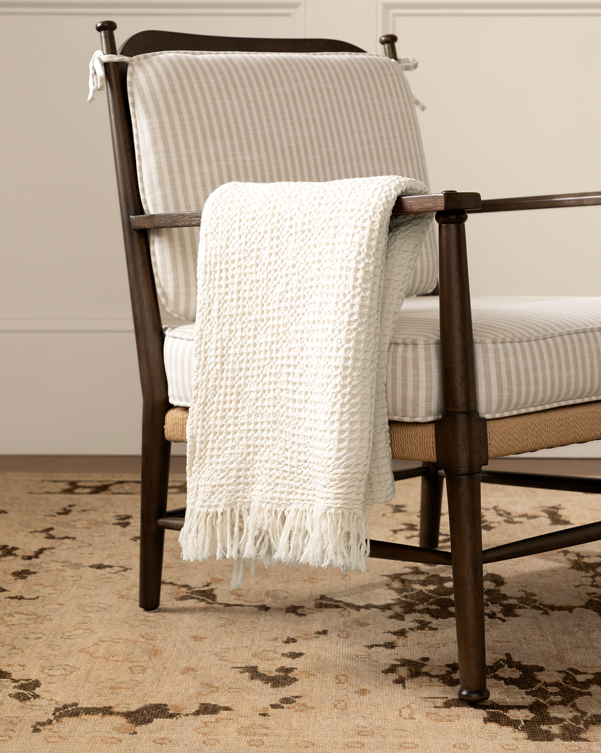 A wooden armchair with light striped cushions sits on a patterned rug. The Viola Throw, an artisanal, textured blanket with fringe, is draped over the chair’s arm, by McGee & Co.