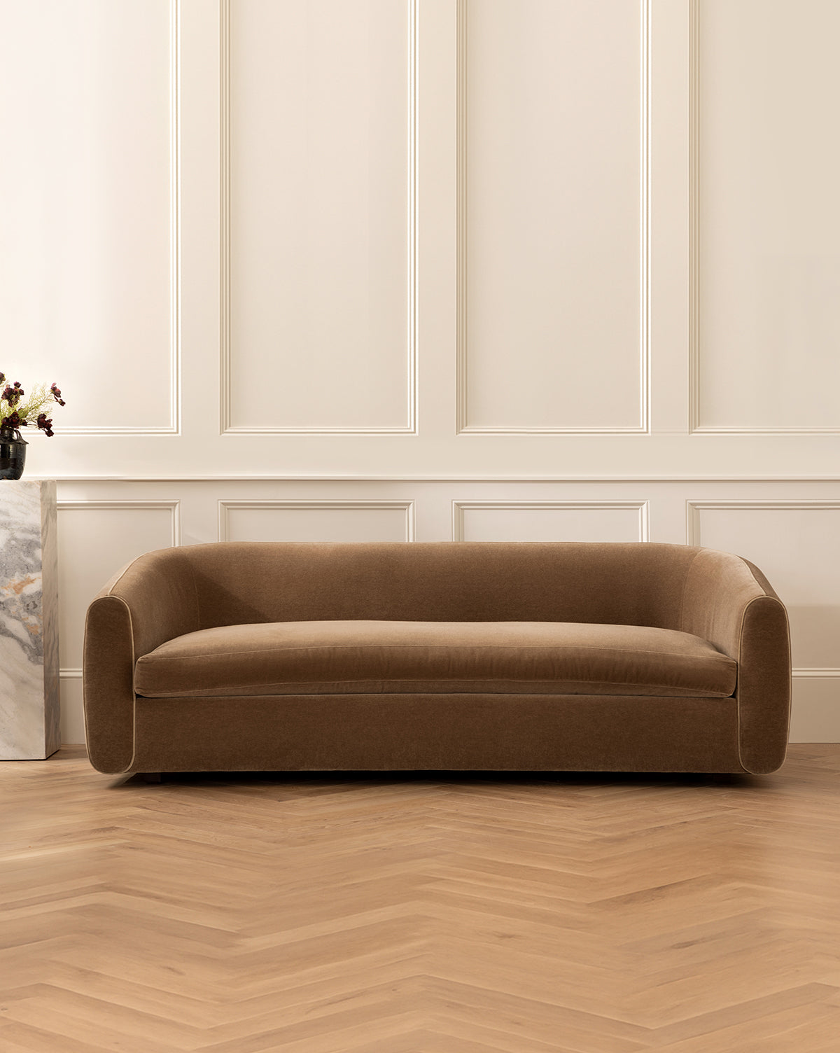 A Wexler Sofa 90 in brown velvet with curved arms sits on a herringbone wood floor against a cream paneled wall. A marble side table with a small vase of flowers is partially visible on the left, by McGee & Co.