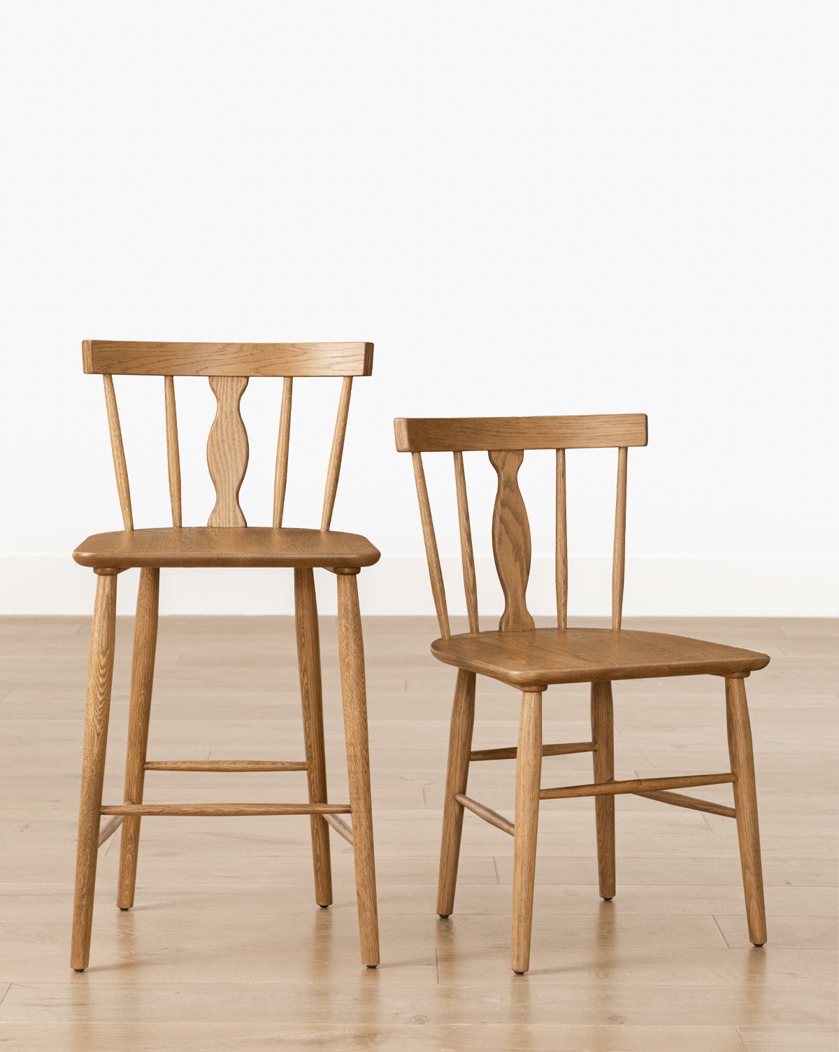 Two Whittaker Chairs—one bar stool and one standard—featuring solid oak construction, spindle backs, and curved seats, are displayed on a light wood floor against a white wall, ideal for elevating any dining space by McGee & Co.