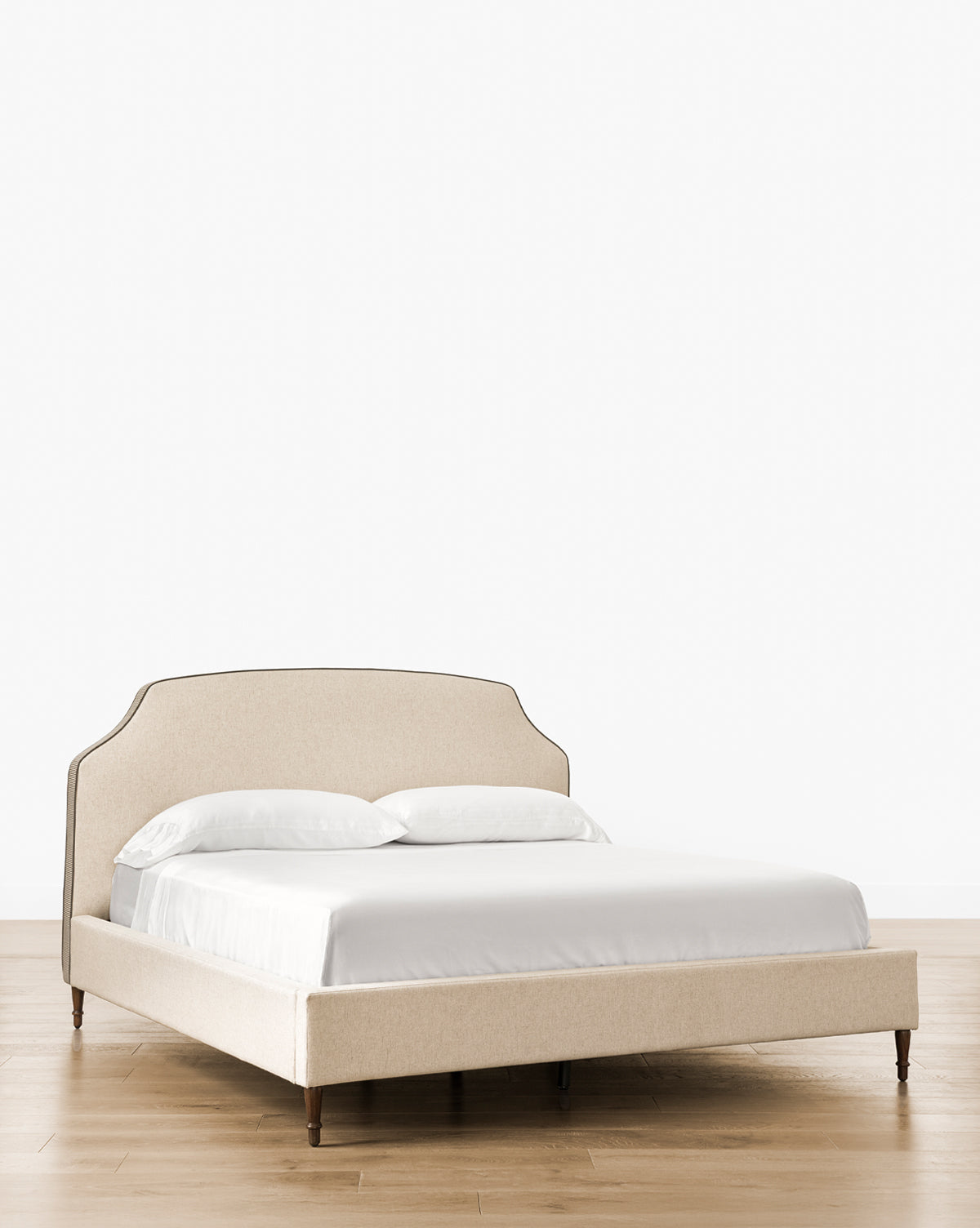 The Willoughby Bed showcases beige linen upholstery, a curved headboard, English Oak legs, white bedding with two pillows, set on wooden flooring against a plain white backdrop, by McGee & Co.