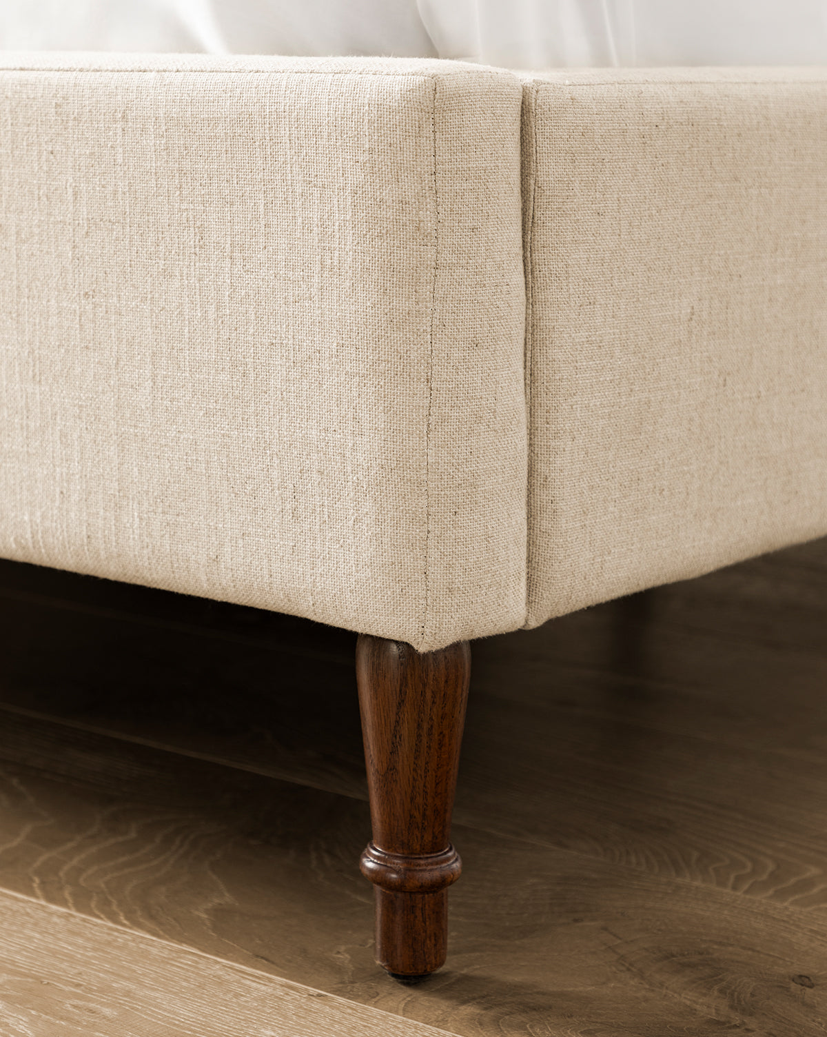 Close-up of the Willoughby Bed’s corner shows its beige linen upholstery and dark English Oak wood legs resting on a hardwood floor. White bedding is partially visible atop the bed frame, by McGee & Co.