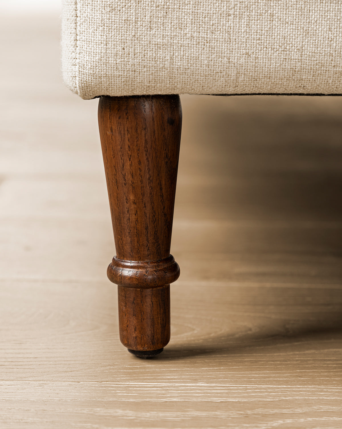Close-up of an English Oak wood leg on the Willoughby Bed's beige linen upholstery, shown resting on a light wooden floor, by McGee & Co.